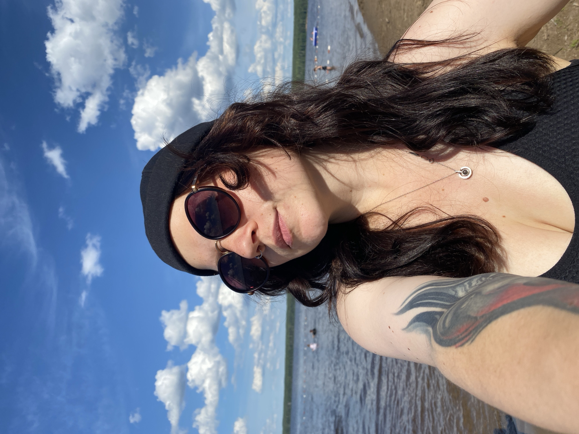 The media file is a vertical, medium-shot, selfie-style photograph of a person outdoors on a sunny day. The subject is a person with dark, long, wavy hair, wearing a black beanie, sunglasses, and a black top. They have a tattoo on their right arm and are wearing a necklace. The person is smiling slightly and looking towards the camera.
The setting appears to be a lake or large body of water, with a sandy or muddy shore visible in the background. The water is calm and reflects the blue sky. In the distance, there are several people in the water, some appearing to be swimming or on inflatable objects. The sky is bright blue with scattered white, fluffy clouds, indicating fair weather and likely midday.
The overall mood of the image is relaxed and summery. The bright sunlight and the presence of people enjoying recreational activities by the water suggest a leisure setting. The location is identified as Chaikovsky, Russia. There is no visible text in the image.