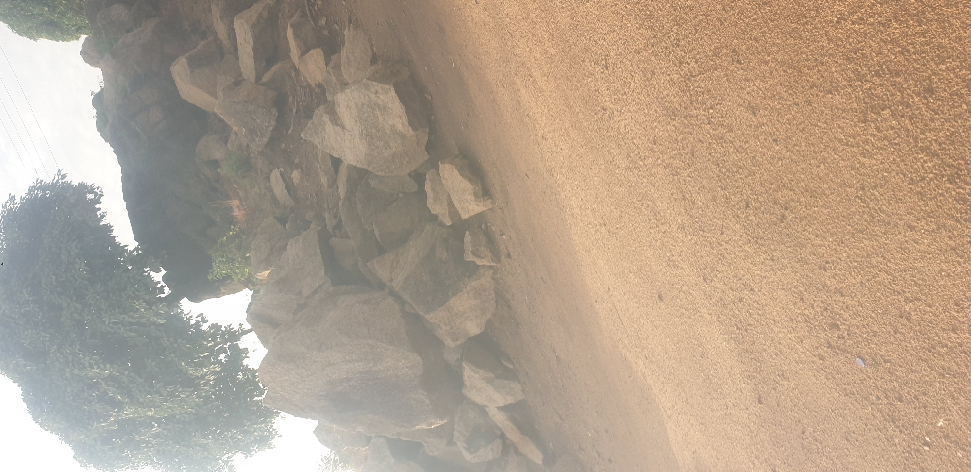 The image depicts a rocky, uneven terrain with large boulders scattered across the scene. A large tree with dense foliage is visible in the upper left portion of the frame, partially obscuring a rock formation. The ground is composed of dirt and sand, with some smaller stones and debris. The overall lighting suggests it is daytime, with sunlight casting shadows and highlighting the textures of the rocks and ground. The environment appears natural and undeveloped. It is not possible to determine the specific location within the city of Jos, Nigeria, or the presence of any people or specific actions from this image alone.