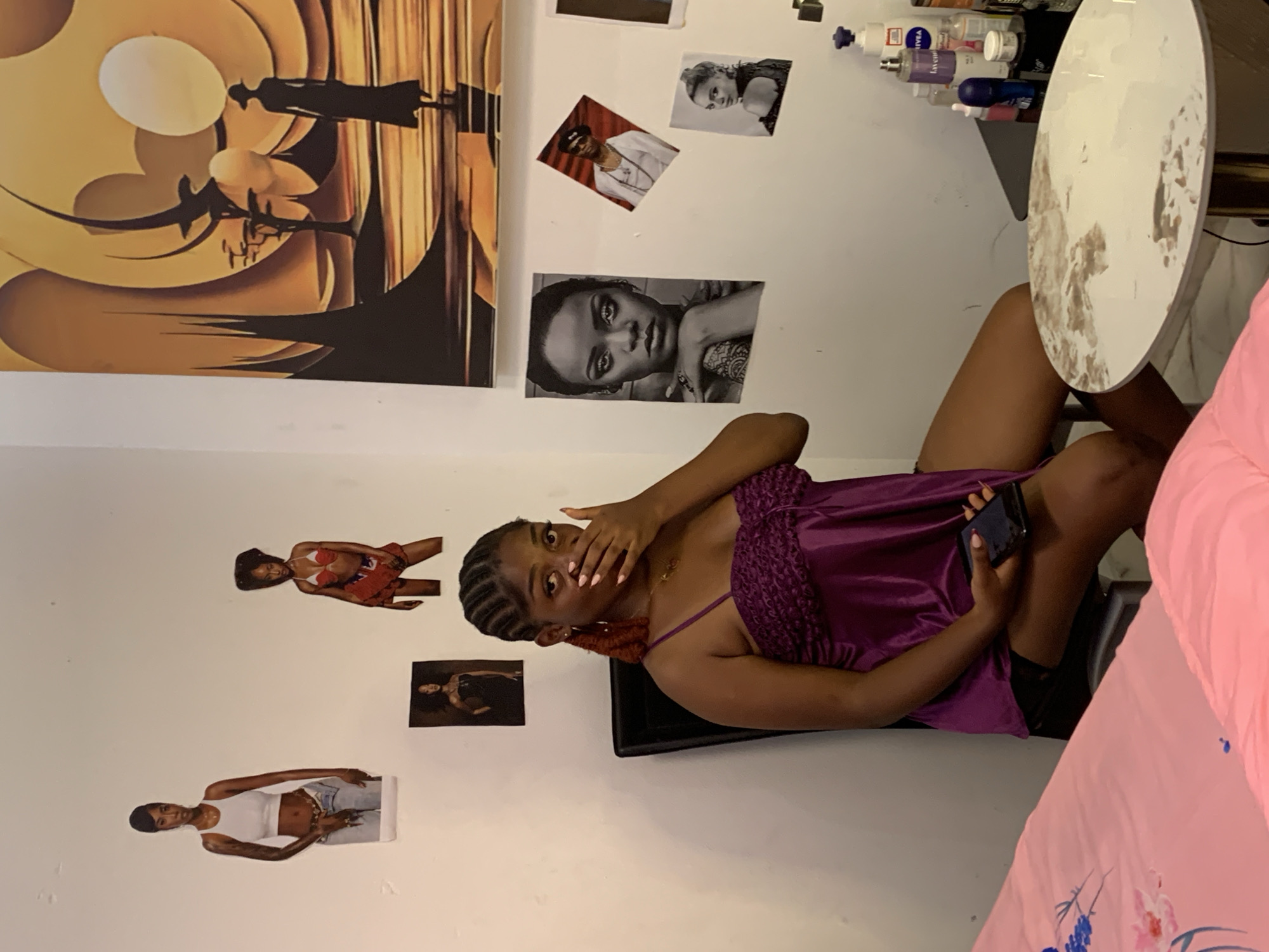 An indoor residential or private setting, located in Lagos, Nigeria, features a young woman seated on a dark, high-backed chair. The woman has dark skin, braided hair with reddish-brown extensions, and is wearing a purple, satin-like spaghetti-strap top with a textured bustline. Her right hand covers her mouth, while her left hand holds a dark-colored smartphone resting on her lap. She wears a gold-colored pendant necklace and a small earring in her visible left ear; her fingernails are painted a light pink or nude color.
The white wall behind the woman is decorated with several cutout images and a large framed artwork. On the left section of the wall, three images are affixed: a woman in a white crop top and jeans (identified as Tiwa Savage), a woman in a red and white bikini top and red shorts, and a smaller, partially obscured image of a woman in a dark dress. The upper right wall displays a large, stylized artwork featuring a warm, golden-orange landscape with abstract circular elements, pointed structures, and the silhouette of a woman in a hat and long dress walking away. Below this artwork, on the right wall, are three additional cutout images: a black and white portrait of Rihanna, a smaller, angled image of a man in a white top and dark hat, and a smaller image of a woman looking over her left shoulder.
In the lower left foreground, a bed with pink bedding, including a floral-patterned sheet with blue and pink flowers, is partially visible. To the woman's right, a dark, small table or shelf holds several bottles and containers, including a tall white Nivea lotion bottle, a tall purple bottle, and a blue roll-on deodorant. A round, white table with a visibly stained or discolored surface and a gold-colored pedestal base occupies the lower right foreground. The scene is illuminated by artificial light.