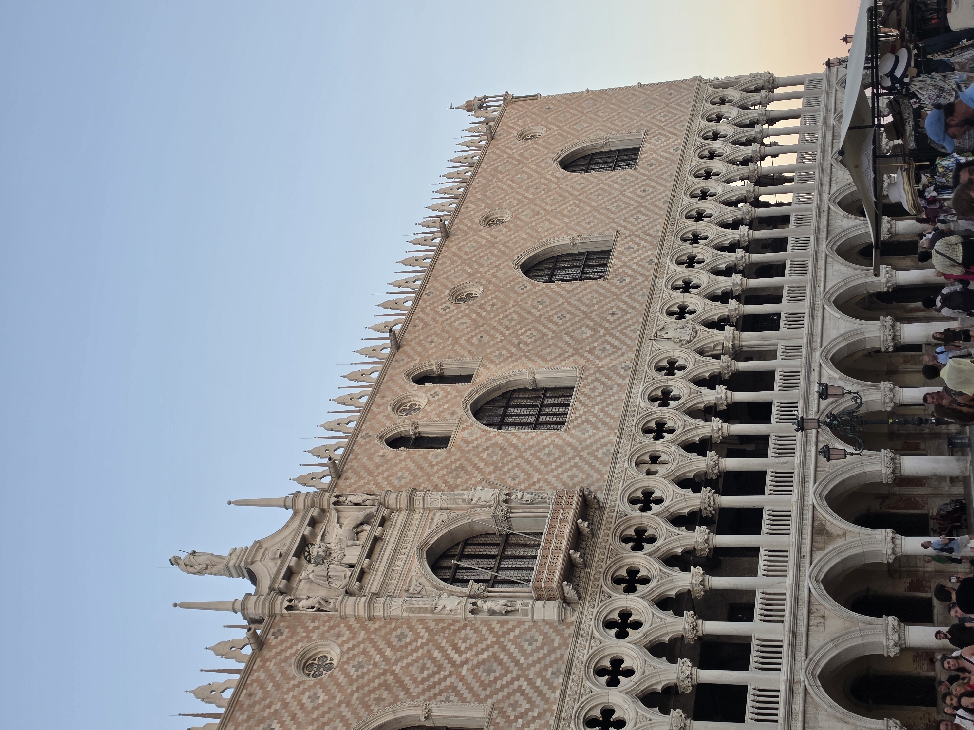 The image captures a close-up view of the Doge's Palace in Venice, Italy, a historic landmark situated in St. Mark's Square. The architecture is characterized by its Gothic style, featuring intricate details such as pointed arches, ornate tracery, and a distinctive diamond-patterned facade. The building is bathed in the warm, soft light of either early morning or late afternoon, casting subtle shadows and highlighting the textures of the stone.
In the foreground, a bustling scene unfolds with several people milling about. They appear to be tourists, some are walking, while others are standing and looking around. A market stall selling various items, including hats and clothing, is partially visible, adding to the vibrant atmosphere of the piazza. The sky above is a clear, pale blue, suggesting fair weather. The overall impression is one of historical grandeur and lively public activity.
