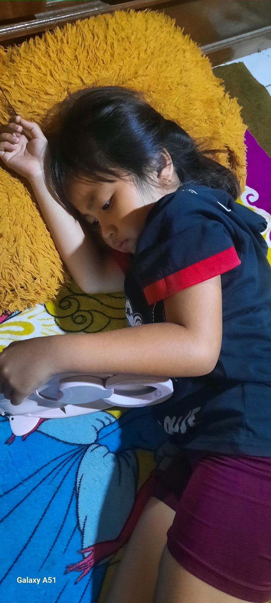 A young girl with dark hair lies on her side on a yellow, fluffy pillow. She is wearing a dark t-shirt with red trim on the sleeves and maroon shorts. Her left arm is bent and resting on the pillow, while her right arm is extended downward, holding a white object with grey accents. The background consists of a colorful blanket with a cartoon character print and a purple fabric. The scene appears to be indoors. The location of the photo is Jatitengah, Indonesia.