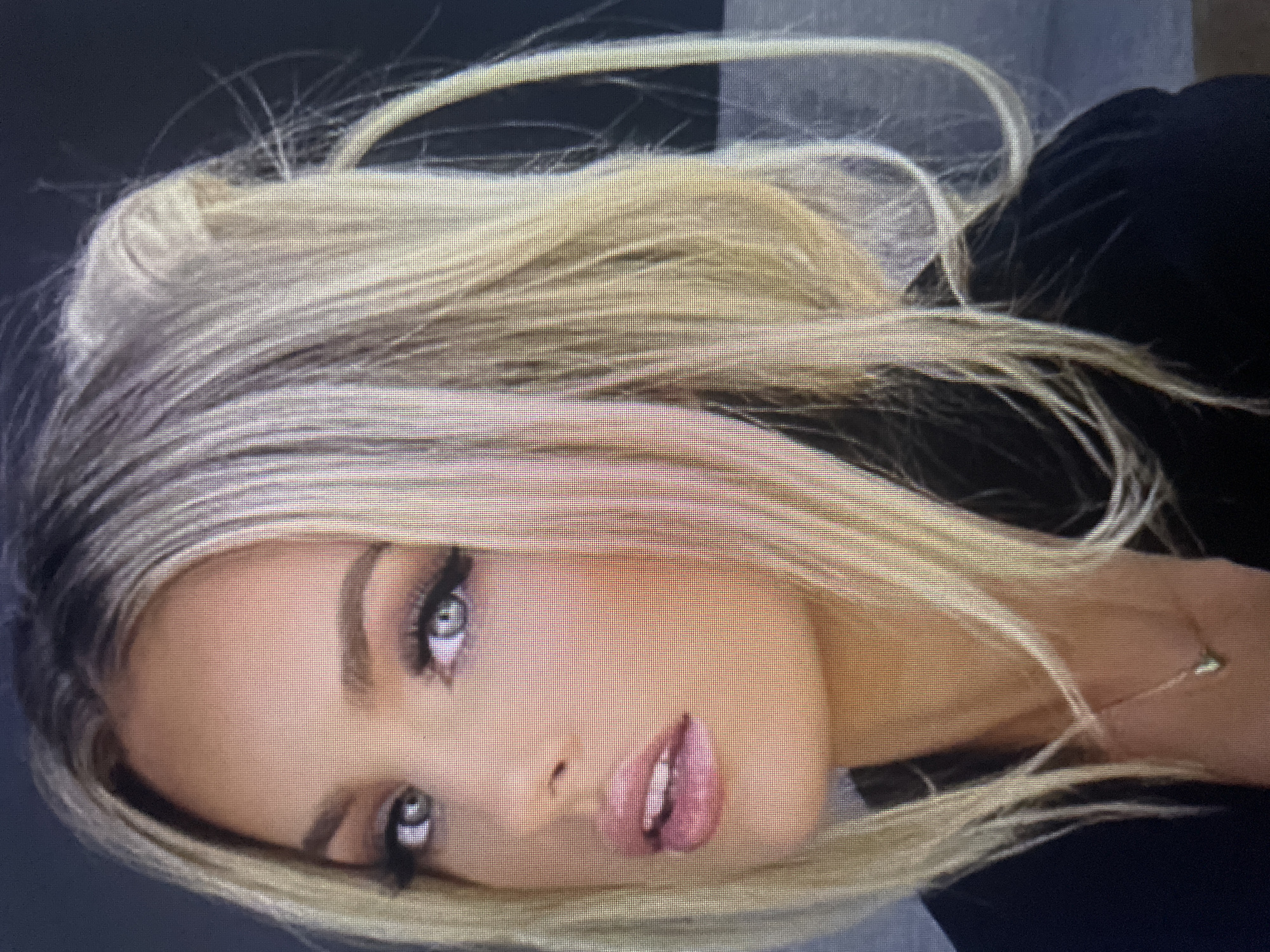 The image is a close-up portrait of a blonde woman with fair skin, gray eyes, and full, pink lips. She is wearing makeup, including eyeshadow and lipstick. Her hair is styled in a messy updo with strands falling around her face. She is wearing a black top and a delicate gold necklace with a small pendant. The background is dark and out of focus. The image does not contain any elements that would allow for the determination of its location in the city of Kołobrzeg, Poland.