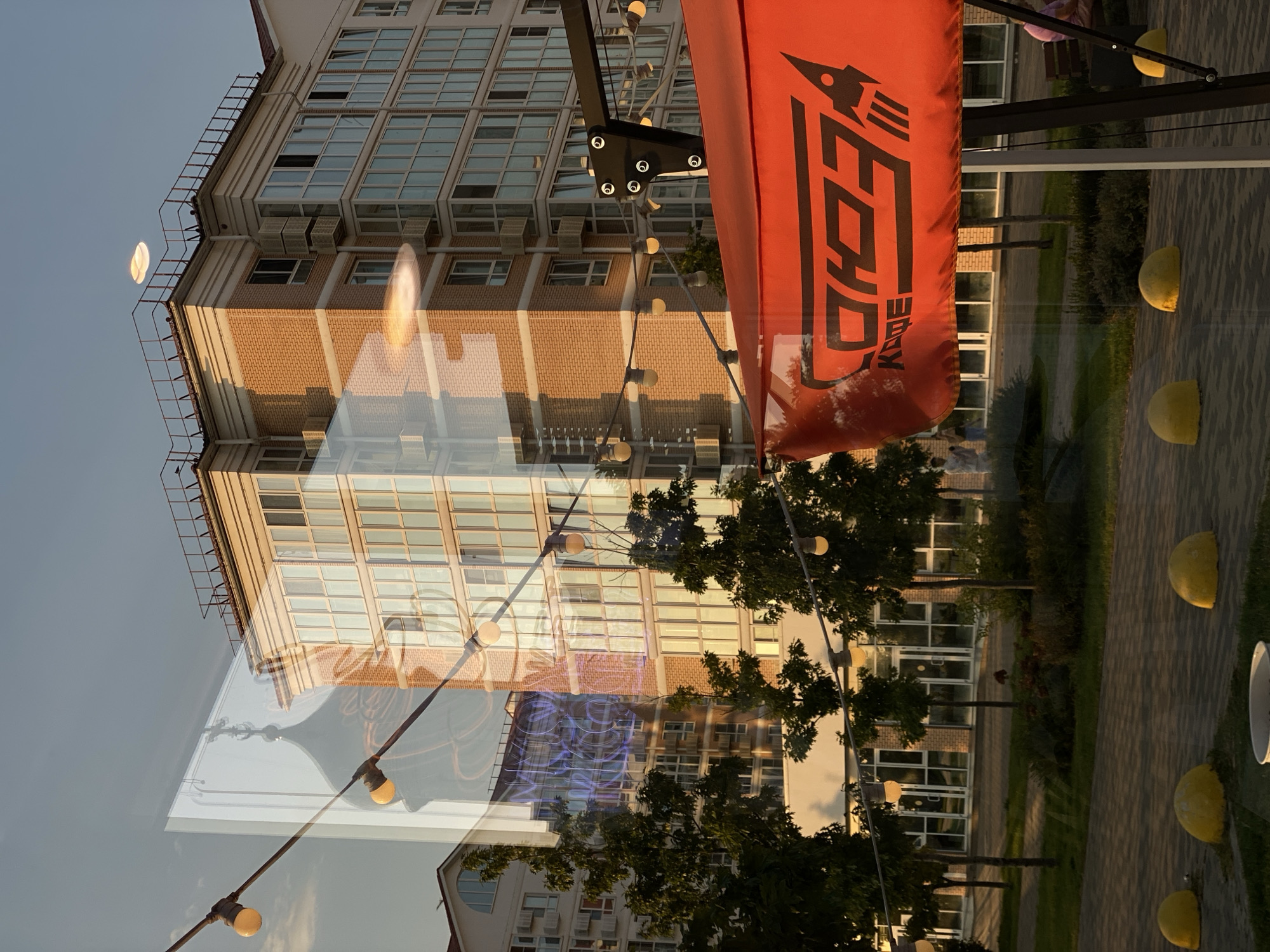 The image captures a reflection on a glass surface, showing a multi-story building with a brick facade and numerous windows. In the foreground, an orange canopy with a stylized logo and the word "КОФЕ" (coffee in Russian) is visible, attached to a black metal frame. Strings of light bulbs are strung across the scene. The reflection on the glass shows a cityscape that includes a domed structure, possibly a church or a historic building, overlaid with neon blue lighting. The overall environment appears to be an urban outdoor space, possibly a cafe or a public area, with trees, green spaces, and paved walkways. The location is identified as Agronom, Russia.