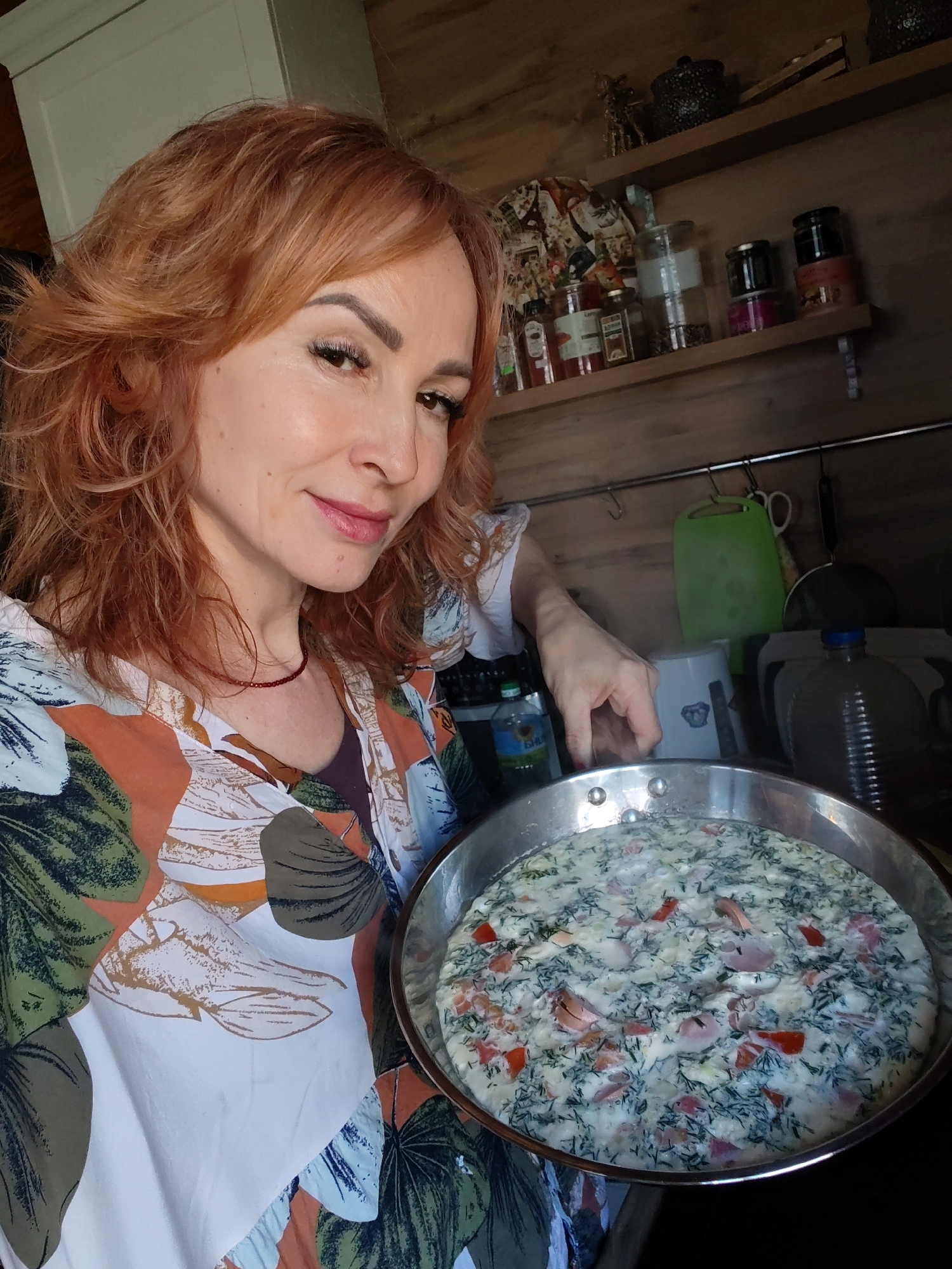 A woman with red hair is holding a large, round stainless steel pan filled with a white liquid dish containing chopped red tomatoes and green herbs, possibly dill. She is smiling and wearing a patterned shirt with white, orange, and green floral designs. Behind her are wooden walls, shelves, and various kitchen items including jars, a kettle, a green cutting board, and a water bottle. The location is Старое Михайловское, Russia.