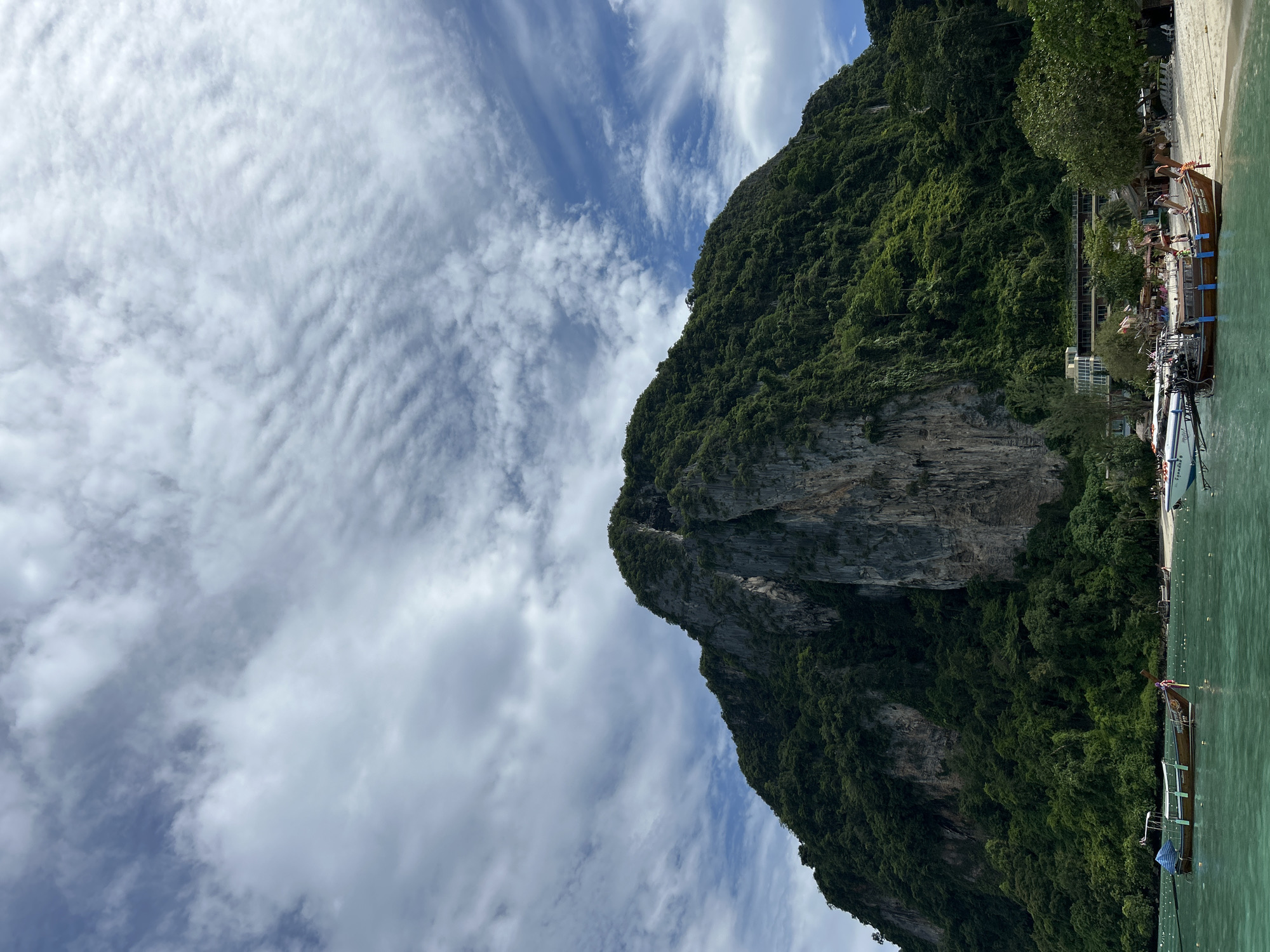 The image captures a scenic view of a large, verdant mountain rising from the sea in Ao Nang, Thailand. The mountain's rocky face is partially visible, with dense green foliage covering much of its surface. The sea in the foreground is a vibrant turquoise color, with several longtail boats moored near the shore. One boat, a traditional wooden design with a striped canopy, is prominently featured in the center left of the image. The beach in the background appears sandy, with some buildings and palm trees visible. The sky above is a bright blue, dotted with wispy white clouds that create a dynamic texture. A few people are visible on the beach and on the boats, appearing to be engaged in leisure activities. The overall impression is one of a tropical paradise, with the majestic mountain serving as a dramatic backdrop to the tranquil coastal scene.