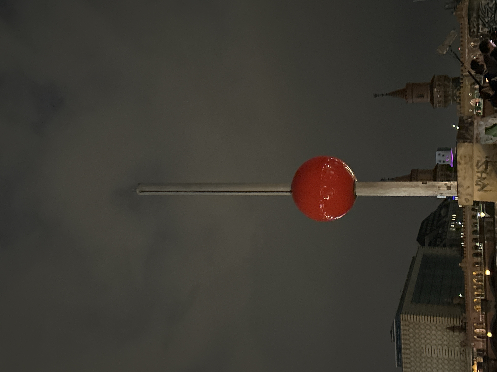 A large, glossy red sphere, possibly an art installation or balloon, is prominently featured in the center, suspended by a slender, light-colored pole against a dark, overcast night sky. The sphere reflects light, giving it a wet or highly reflective appearance.
In the background, an urban landscape unfolds at night. To the right of the sphere, a distinct brown brick tower with a pointed roof, resembling part of a historic bridge or gate, is visible. Further to the right, outlines of people can be seen, suggesting an audience or passersby.
Below the sphere and to its right, modern buildings with illuminated windows and complex facades are visible. One building exhibits a distinct pattern on its surface and the numerical sequence "10000000" clearly displayed. Adjacent to this, on a wall or structure, faint text, including the word "SHAN," appears to be graffiti.
The scene takes place at night, as indicated by the dark sky and the artificial lights emanating from the buildings. The overall setting appears to be an urban area, likely a public space featuring an illuminated art piece or landmark.