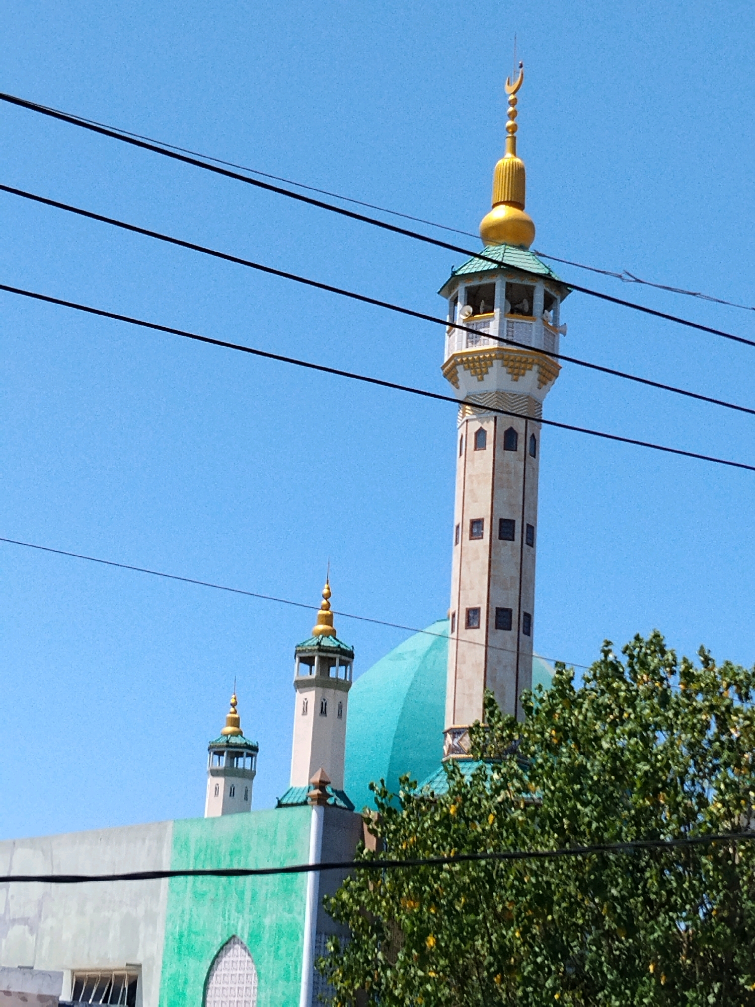 This is a photograph taken from a low angle, looking up at a mosque with multiple minarets and a large dome. The mosque appears to be located in an urban setting in Surabaya, Indonesia, as indicated by the presence of power lines crisscrossing the sky in the foreground, a common sight in Indonesian cities.
The most prominent feature is the tallest minaret, which is light-colored with dark rectangular windows arranged vertically. It is topped with a green roof and a golden spire crowned with a crescent moon. To the left of this main minaret, there are two smaller, similar minarets, also topped with green roofs and golden spires.
A large, turquoise dome is visible behind the minarets and the mosque's building structure. The building itself has a section painted in a light grey, and another section in a vibrant green, featuring an arched window with a decorative lattice. A dense green tree with some yellow foliage partially obscures the lower portion of the mosque and the right side of the image. The sky is a clear, bright blue.
There are no people visible in the photograph. The focus is solely on the architecture of the mosque and its immediate surroundings, with the power lines serving as a contextual element of its urban placement. The overall impression is of a bright, sunny day.