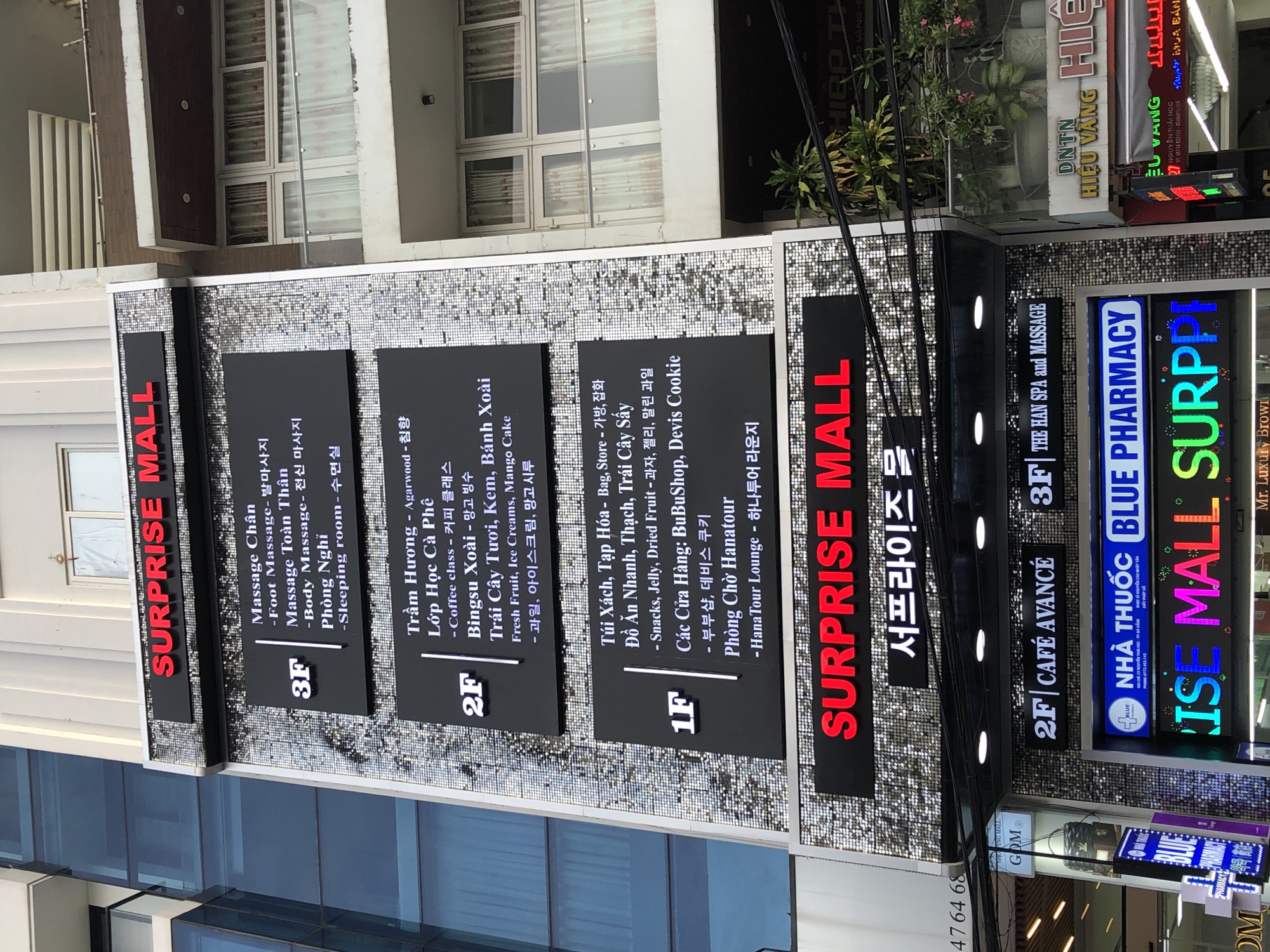 The image shows a multi-story building with prominent signage in Đà Nẵng, Vietnam. The building features a facade with a reflective, mosaic-like pattern. Several businesses are advertised on the building's exterior.
At the top, a large red neon sign reads "SURPRISE MALL". Below this, on a black rectangular panel, the third floor (3F) lists "Massage Chân", "Foot Massage", "Massage Toàn Thân", "Body Massage", and "Phòng Nghỉ" with Korean text.
Further down, the second floor (2F) is also advertised on a black panel. It lists "Trầm Hương - Agarwood", "Lớp Học Cà Phê", "Coffee class", "Bingsu", "Trái Cây Tươi, Kem, Bánh Xoài", "Fresh Fruit, Ice Creams, Mango Cake", and associated Korean text.
The first floor (1F) displays "Túi Xách, Tạp Hóa - Bag, Store", "Đồ Ăn Nhanh, Thạch, Trái Cây Sấy", "Snacks, Jelly, Dried Fruit", "Các Cửa Hàng: BuBuShop, Devis Cookie", "부부샵, 데비스 쿠키", and "Phòng Chờ Hanatour - HanaTour Lounge".
Near the bottom of the building, another "SURPRISE MALL" sign is visible, this time with multi-colored neon lights. A smaller sign below it displays Korean text.
Further down, at ground level, a blue neon sign reads "2F CAFE AVANCÉ". To its right, another sign says "2F THE HAN SPA and MASSAGE". Below these, a prominent blue and white sign states "BLUE PHARMACY" with Vietnamese text and a phone number.
To the right of the building, the facade has windows with curtains, suggesting interior spaces. Other businesses are visible in the background, including one with a yellow sign reading "HIỆU VANG" and another with "DNTN". The environment appears to be a street scene with power lines and some vegetation visible. No people are clearly discernible in the image.