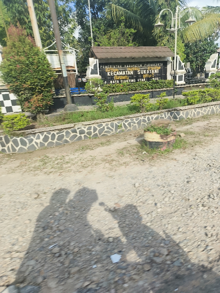 The image shows a sign in front of a building in Sukatani, Indonesia at the coordinates provided. The sign, which is in Indonesian, reads "Pemerintah Kabupaten Furwakarta Kecamatan Sukatani Jl. Raya Cianting Utara". There is a stone wall with a small patch of green grass in front of the sign. The ground is gravel. There are three indistinct shadows of people in the foreground, appearing to be walking away from the sign. The shadows are blurry. There is a tall metal pole behind the sign with two streetlights at the top. In the background, there are trees and a metal fence. The sky is bright blue with some clouds.