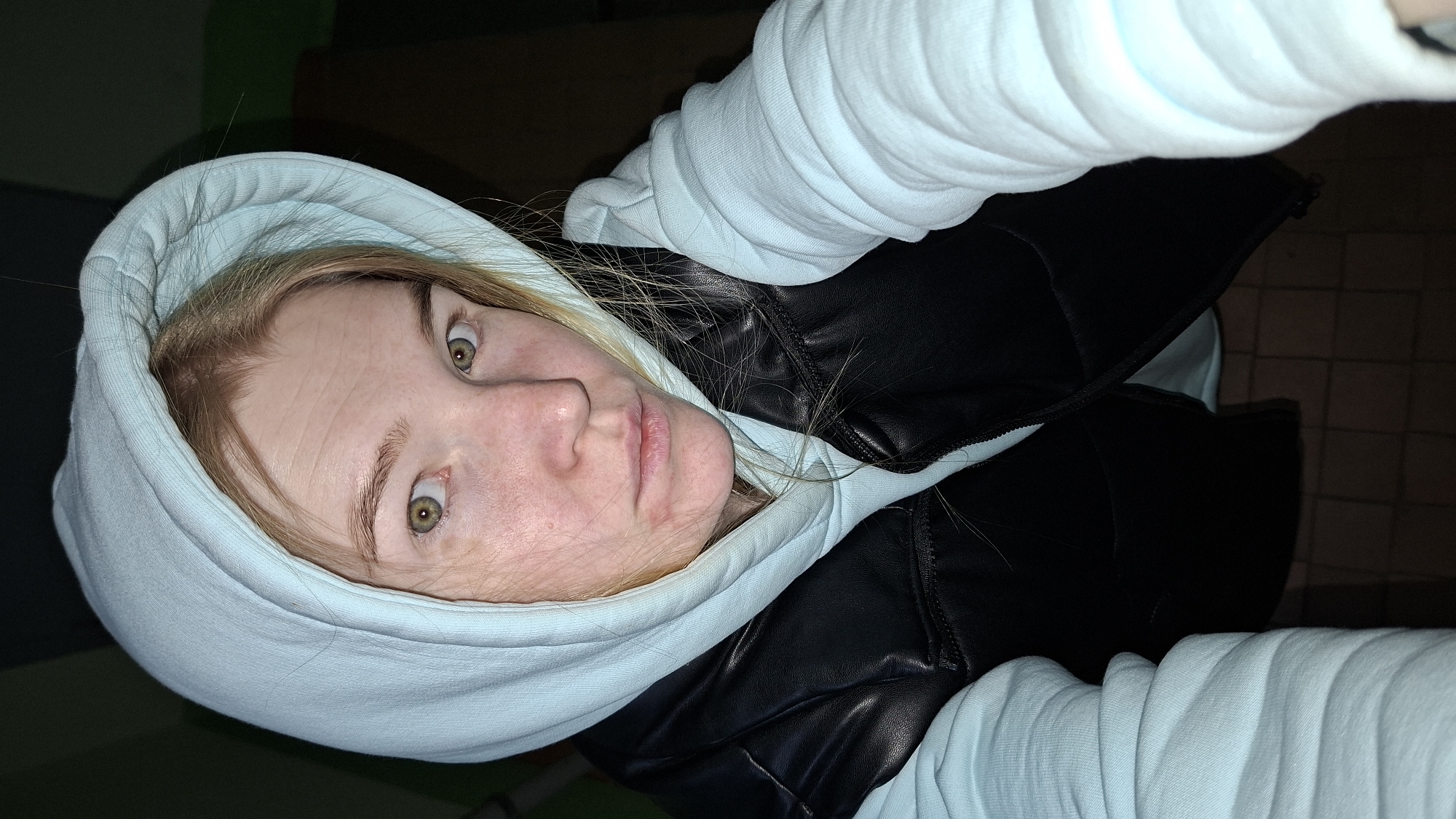 A young woman with light blonde hair and green eyes is posing for a selfie. She is wearing a light blue hooded sweatshirt with a black, quilted vest over it. The photo appears to be taken at night. The location may be Chaikovsky, Russia, based on the prompt. The background is dark and includes some hints of interior structure with green elements.
