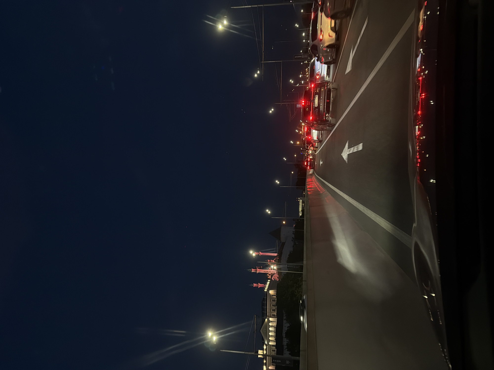 The image shows a nighttime scene in Kazan, Russia. The view is likely from a bridge, with a road leading forward. The road is marked with white lines and a large arrow indicating the direction of traffic. There are multiple cars on the road, with their red taillights visible, suggesting they are stopped or moving slowly. Streetlights illuminate the road and the surrounding area. In the distance, illuminated buildings are visible, including structures with distinctive towers, suggesting the presence of historical or cultural landmarks. The sky is dark, with few visible stars. The right side of the image includes the side of a vehicle, potentially the one from which the photo was taken.