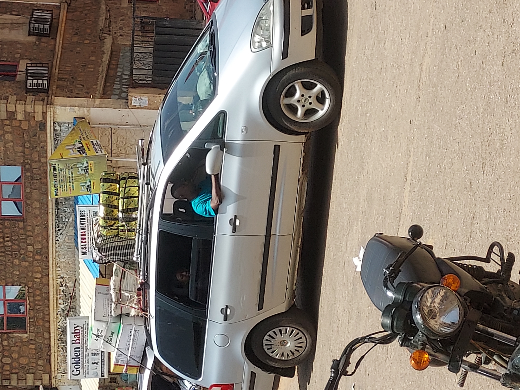 A silver minivan overloaded with goods is parked on a street in Jos, Nigeria. The minivan is stacked with boxes labeled "Golden Baby," "Adcor," and "AoToy," along with sacks of unidentified materials. A sign reading "MUSA CHINA VENTURES" is visible near the boxes. A man wearing a blue shirt is in the driver's seat. A motorcycle is parked next to the minivan. The buildings in the background are made of brown stone with red-framed windows.