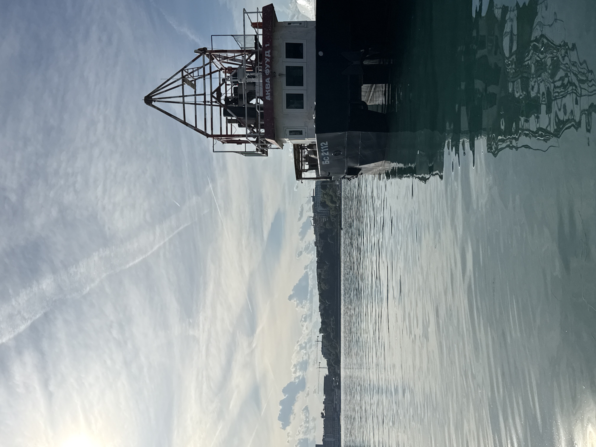 The image shows a vessel named "АКВА ФУУД 1" with registration number "БС 2112" docked in the city of Nesebar, Bulgaria. The vessel is partially obscured by its reflection on the water. The sky is visible in the background. A landmass with buildings and trees is visible on the horizon behind the boat. The boat has a metal framework on its top.