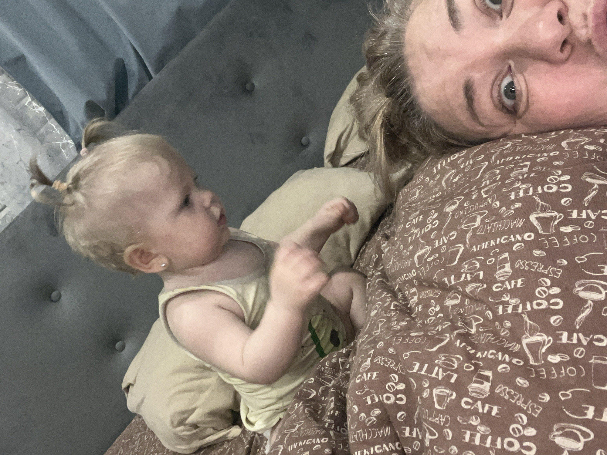 A woman and a baby are lying on a bed. The woman, with graying blonde hair, is on her side facing the camera with her head resting on a pillow. A brown comforter with white coffee-themed text and images covers the bed. Next to her, a baby with blonde hair styled in small pigtails is lying on a light yellow pillow, facing away from the camera. The baby is wearing a light yellow tank top. The background includes a gray upholstered headboard and a portion of a curtain. The location is likely indoors in Vesyoloye, Russia.