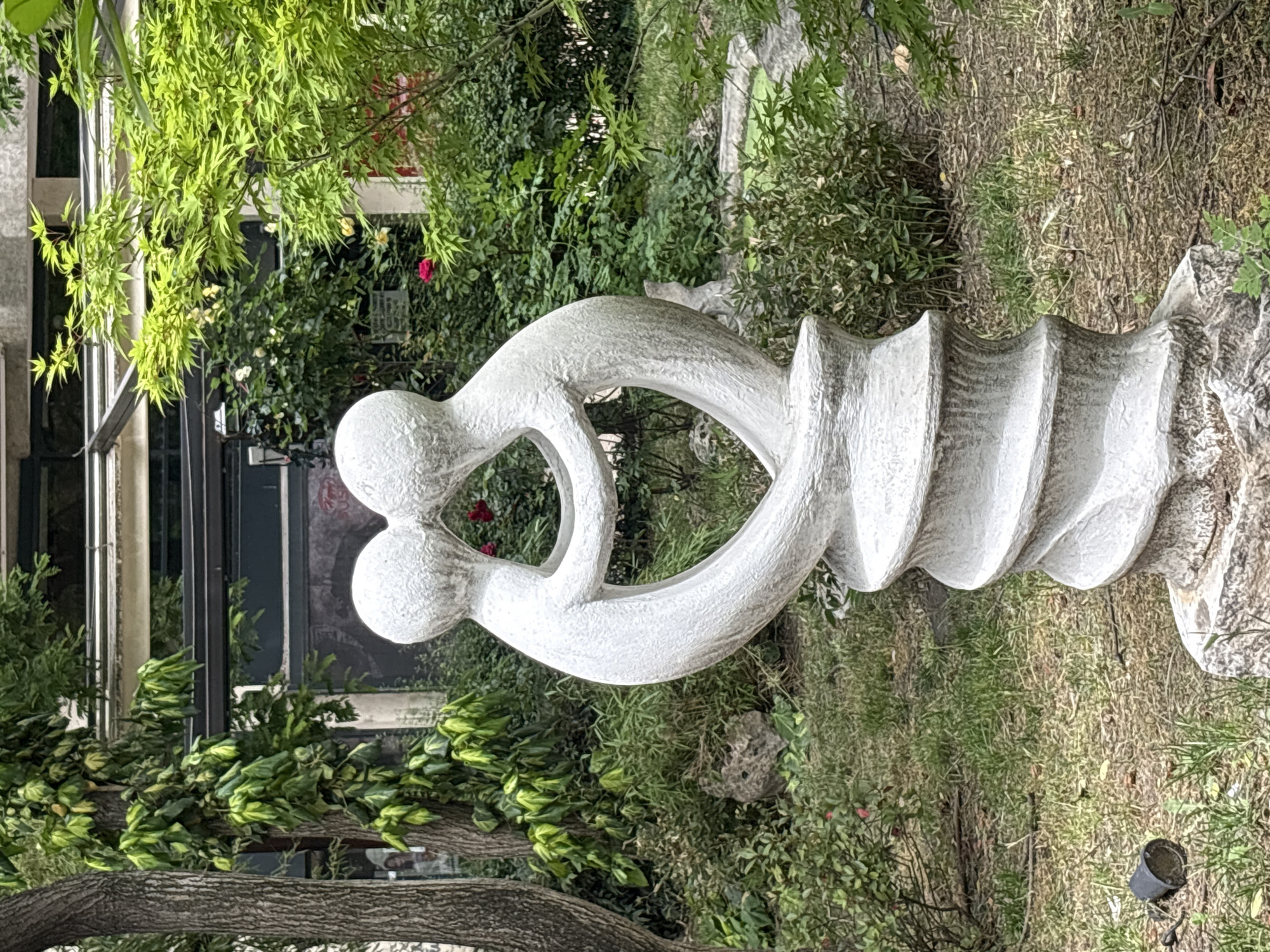 The image features a white, abstract sculpture in a garden setting. The sculpture appears to depict two figures with interconnected heads, forming a heart-shaped opening, adjoined to a vertically aligned, textured structure resembling a screw thread. The background shows dense green vegetation, including trees and shrubs. Behind the vegetation, a building with windows is visible. The ground is covered with grass and patches of dirt or dead leaves.
Based on the characteristics of the sculpture and its setting, the location is likely in or near the Old Town of Plovdiv, Bulgaria, known for its gardens and art installations.