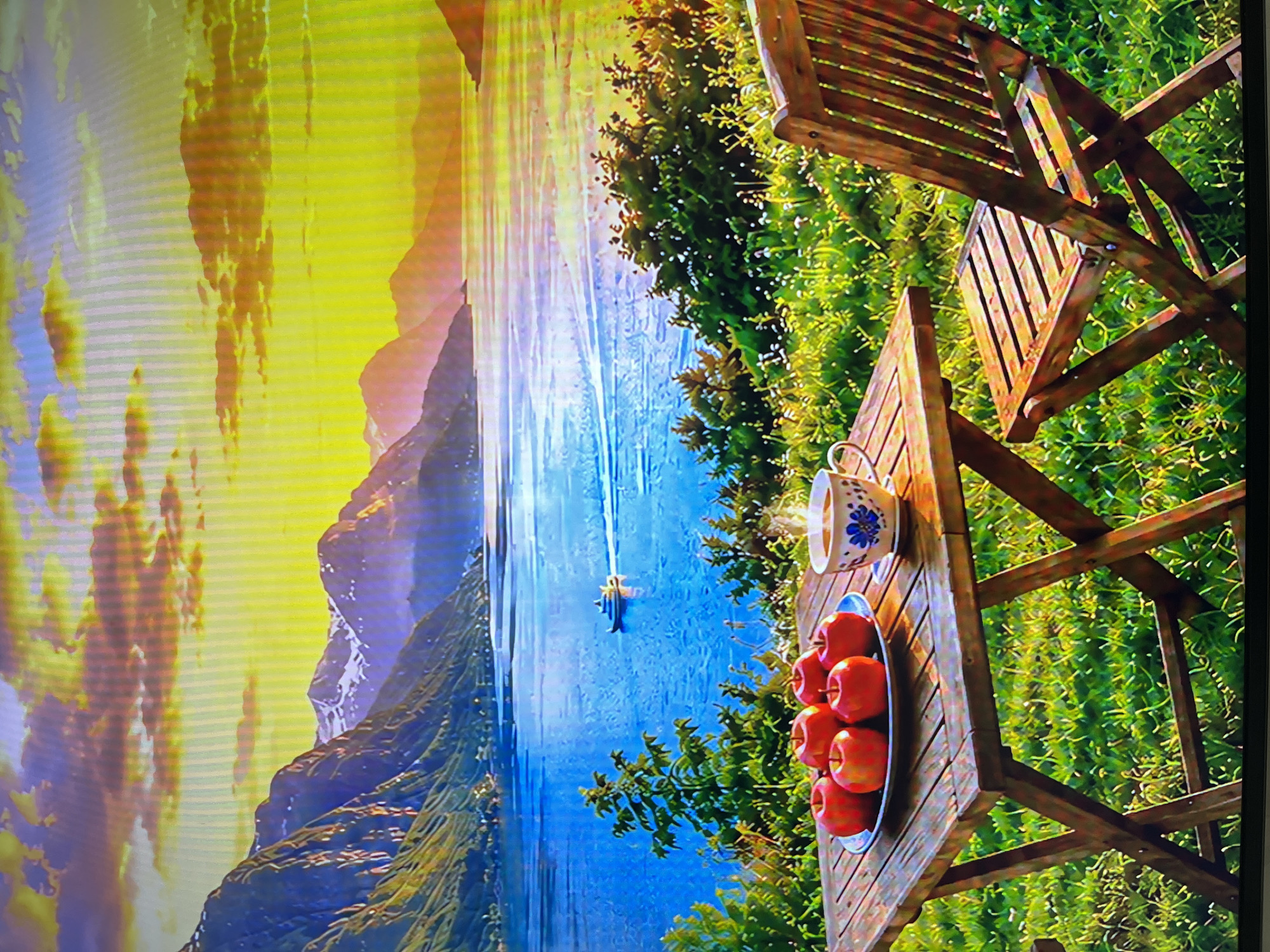 The image depicts a serene outdoor scene with a table and chair set on a grassy area overlooking a body of water and mountains. On the table, there is a plate of five red apples and a steaming cup of coffee. In the background, a boat sails on the blue water, with a backdrop of dramatic mountains under a vibrant, sunset sky. The environment is lush with green grass and foliage. This scene is not located in the city of Kharkiv, Ukraine.
