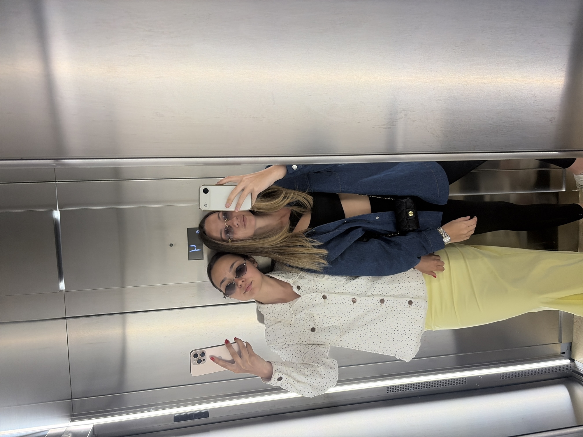 Two women are reflected in a mirrored elevator. One woman is wearing a white shirt with small dots, sunglasses, and a yellow skirt. She is holding a pink phone. The other woman is wearing a black top, black pants, and a blue jacket. She is holding a white phone. The elevator floor is visible at the bottom. The elevator button panel is at the top left.