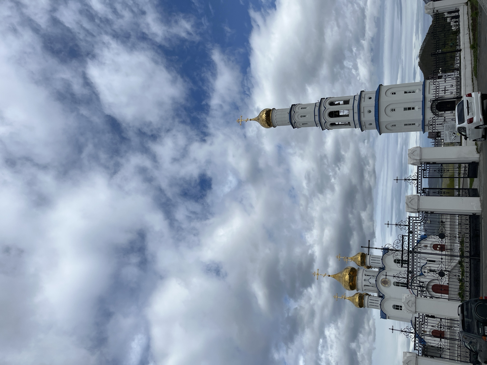 The image depicts the Ascension Cathedral, a prominent Orthodox Christian church located in Petropavlovsk-Kamchatsky, Russia. The cathedral, distinguished by its white facade and multiple golden domes topped with crosses, stands against a backdrop of a dynamic sky filled with dramatic clouds. Adjacent to the cathedral is a tall, slender white bell tower, also adorned with a golden cross. The scene is enclosed by an ornate black metal fence with white pillars, and in the foreground, a white sedan is parked near the entrance. The overall impression is one of a solemn and architecturally significant religious site under a partly cloudy sky.