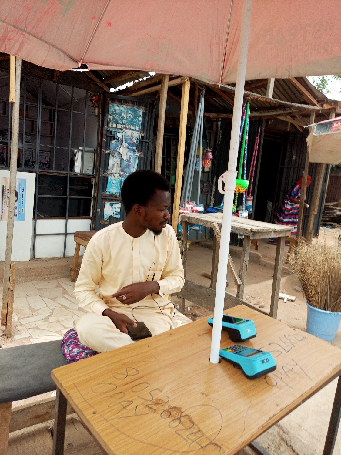 A man wearing a light yellow traditional outfit with earbuds is seated at a wooden table under an umbrella. He is holding a smartphone in his left hand, and his legs are crossed. On the table are two turquoise point-of-sale (POS) devices and what appears to be a scrawled phone number. To the right of the table, there is a blue bucket filled with brooms. Behind the man, there is a storefront with a metal grate and posters on the wall. In the background, other buildings and structures are visible. Based on other available information, the location is Tempure, Nigeria.
