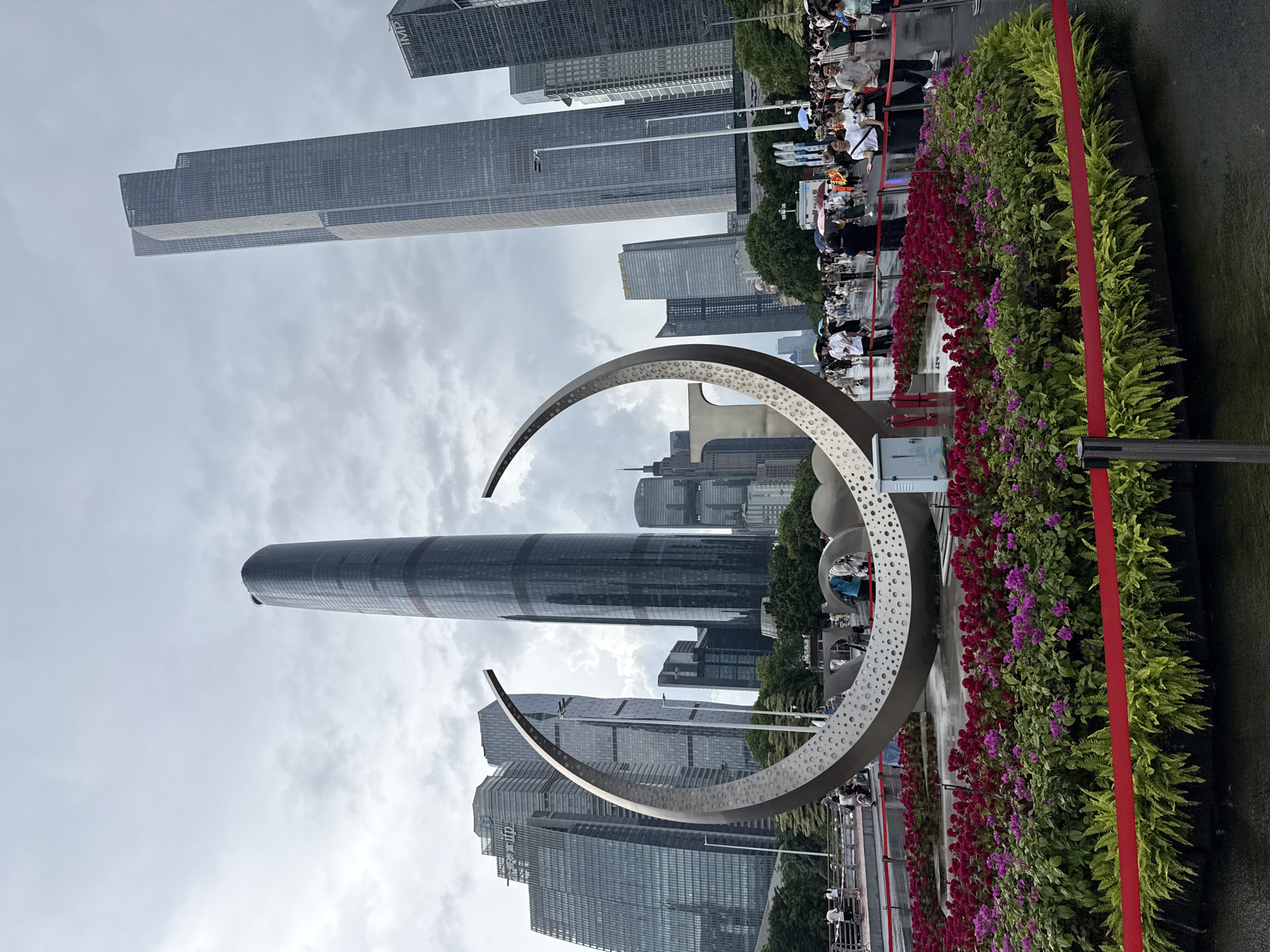 The image depicts a circular flowerbed with red and purple flowers, fronted by a red ribbon barrier, in front of a large, modern metal sculpture. Behind the sculpture and flowerbed, a bustling cityscape of tall skyscrapers rises under a cloudy sky. Numerous people are visible walking and standing around the area, some holding umbrellas, suggesting it might be raining. In the background, the Guangzhou IFC (International Finance Center) and other modern buildings dominate the skyline. The overall scene is of a public urban space, possibly a plaza or a park, within Tianhe District, China, characterized by its impressive architecture and a blend of natural elements with contemporary design.