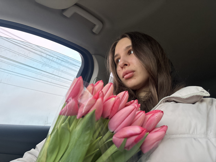 The image captures a young female individual seated as a passenger within a vehicle, observed from a slightly elevated, frontal-left perspective. She possesses long, dark brown hair, fair skin, and full lips, with her gaze directed upward and towards the left, out of the vehicle's window. She is attired in a light-colored, possibly cream or white, puffer jacket with a visible zipper line. Her left arm is partially visible, supporting a substantial bouquet of approximately 15-20 vibrant pink tulips, encased in clear plastic wrapping, which occupies the lower left and central foreground. The tulips exhibit green stems and are tightly clustered. The interior of the vehicle shows a light-colored, possibly beige or grey, overhead grab handle near the window frame. Through the vehicle's side window, an overcast sky is visible, featuring muted blue and white tones, intersected by multiple faint, horizontal lines that may represent overhead wires or reflections. The specific location within the city of Slavyansk-na-Kubani, Russia, cannot be ascertained solely from the visual evidence present in this image.