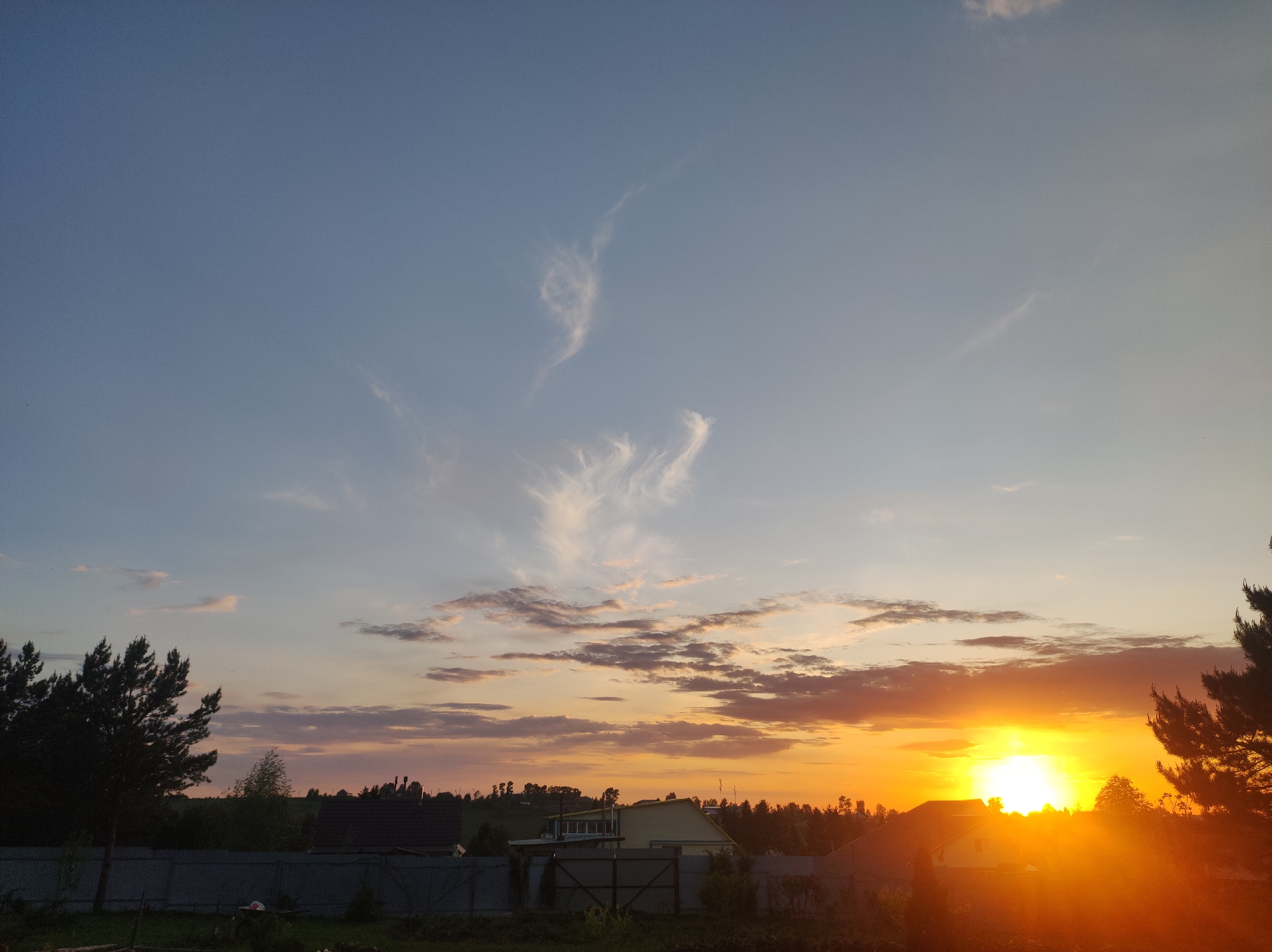 The image depicts a sunset sky over the city of Фоки, Russia. The sky transitions from blue at the top to orange and red near the horizon, with some clouds in the middle section. In the foreground, there's a glimpse of houses and trees silhouetted against the bright light of the setting sun. A fence runs across part of the lower view. The light from the sunset obscures many details of the lower area.