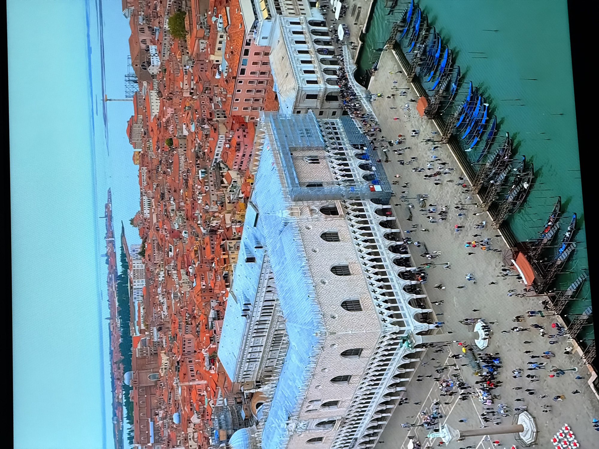 This is a high-angle, aerial view of a bustling city scene, likely in Venice, Italy, given the architectural style and presence of gondolas. The image is vertically oriented, showing a dense cityscape of buildings with terracotta roofs, interspersed with canals.
In the foreground, a large, ornate white building with arched windows and intricate stonework dominates the lower part of the frame. In front of this building is a large open square filled with numerous people walking around, some gathered in groups. To the right of the square, a canal is lined with numerous blue gondolas docked along a pier. More people are visible walking along the waterfront and boarding or disembarking from the gondolas. The water is a vibrant turquoise green.
Further back, the city stretches out with countless red-roofed buildings and more canals. A tall, slender tower or obelisk is visible in the distance. The sky is a pale blue, suggesting daytime with clear or partly cloudy weather. The overall atmosphere appears lively and active, with many tourists and locals present. There is no visible text in the image.