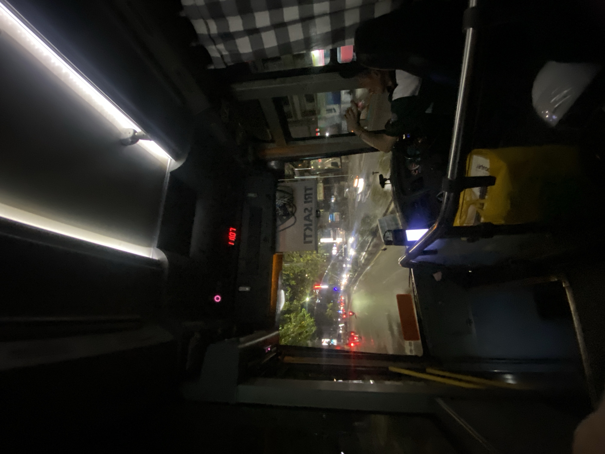 The image is taken from inside a bus at night, looking out the front windshield. The bus is on a city street in Banjarmasin, Indonesia, with traffic visible. The road appears wet, suggesting it may have rained. Trees and buildings line the street. Inside the bus, a digital clock displays "11:07". A driver, or passenger, wearing a dark shirt with a white stripe is visible in the driver's seat, with their arm raised as if holding something. A yellow bag is hanging from a bar in the foreground. The interior of the bus is dimly lit with overhead fluorescent lights.