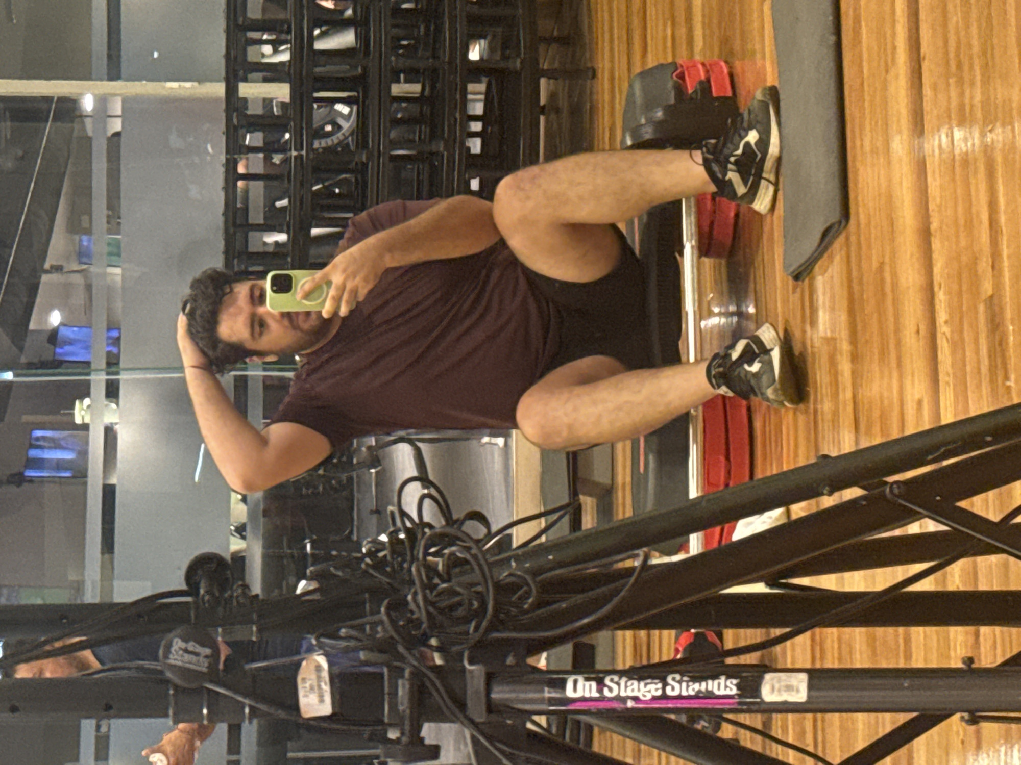 A man is holding a cell phone and taking a selfie while sitting on a bench in a room that appears to be a gym or studio with wood floors. The man is wearing a dark maroon shirt and dark shorts and black-and-white sneakers. He is leaning against a weight set and a black exercise mat. An "On Stage Stands" equipment stand is visible in the foreground. The location is potentially in the city of Puebla, Mexico.