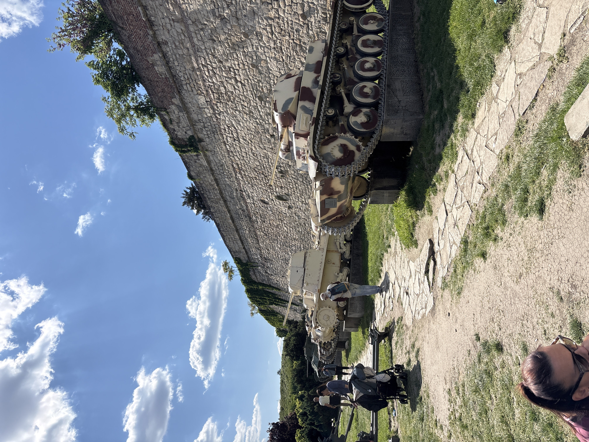 The image shows an outdoor scene in Belgrade, Serbia, likely at the Kalemegdan Fortress complex. A row of military tanks is displayed along a path adjacent to a stone wall. A few people are visible: one man is standing next to a tank; others are in the background near the tanks, and a woman with glasses appears to be in the foreground corner. In the background, there is a blue sky with white clouds and some trees.