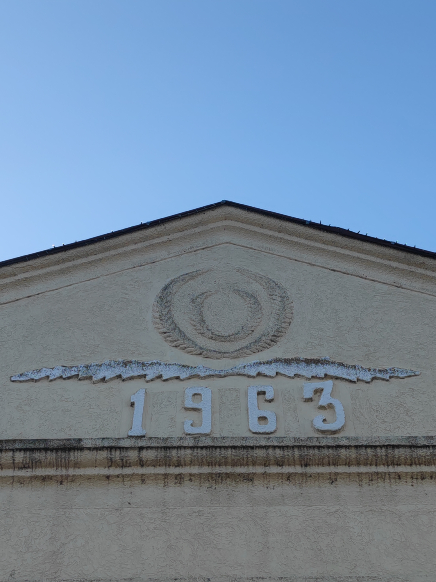 A low-angle, full shot captures the upper portion of a building, specifically the peaked section above the main structure, with the year "1963" prominently displayed. Above the numbers, a decorative element resembling stylized wings or foliage flanks the year. Above this, a circular or wreath-like design is centered within the peak. The building's exterior is textured, and the number and decorative elements appear to be raised or applied to the facade. The photograph is taken during daylight hours under a clear, blue sky. The location of the structure is in Sennoy, Russia.