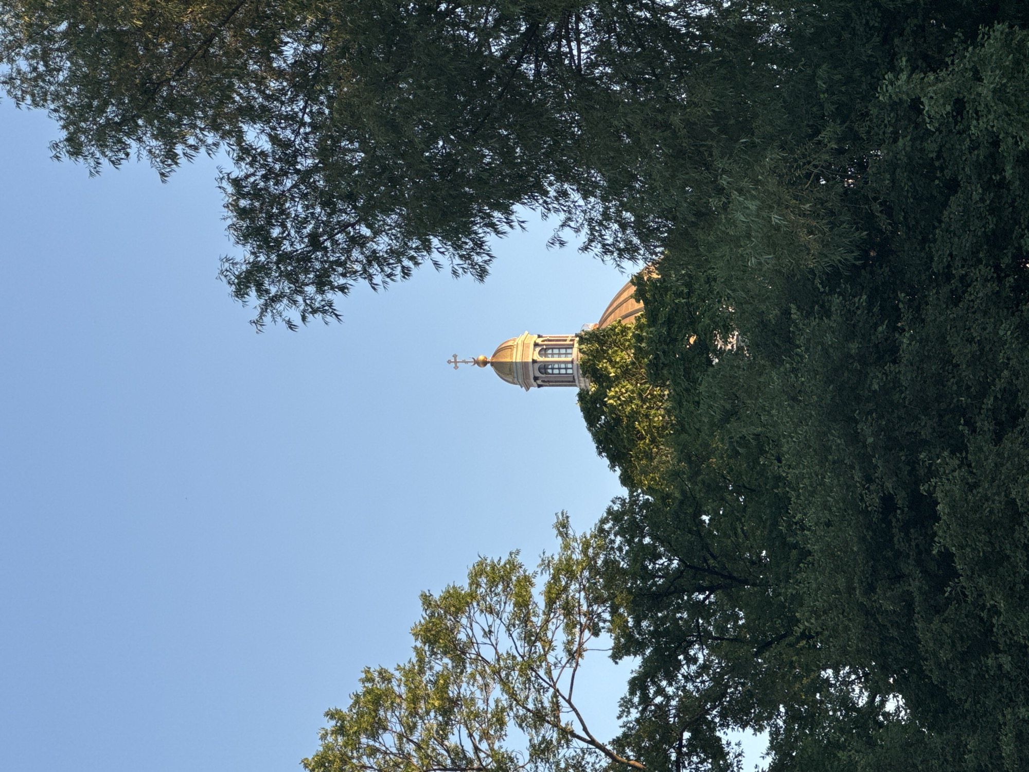 The image shows a golden dome topped with a cross, partially obscured by the lush green foliage of trees. The sky is clear and blue, suggesting it is daytime. The architectural style of the dome, with its classical detailing and lantern, is characteristic of many religious buildings in Saint Petersburg, Russia. While the exact building is not identified, it is likely a prominent landmark within the city, such as a cathedral or a church, given the prevalence of such structures with gilded domes in Saint Petersburg's skyline. The scene is tranquil and serene, with the natural elements of trees framing the religious edifice.