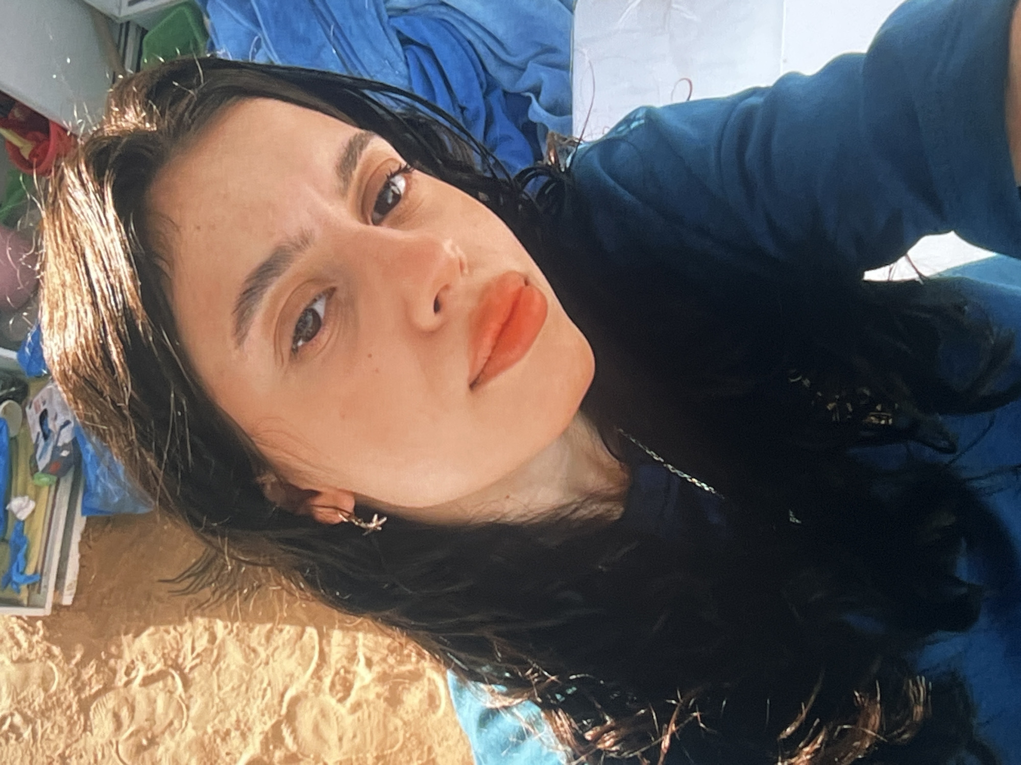 The image shows a close-up, vertically oriented selfie of a young woman lying on sand. She has long, dark hair, dark eyes, and is wearing a dark blue top. Her lips are painted with a bold orange lipstick. She is wearing a small, star-shaped earring in her left ear. In the background, there are folded blue fabrics and some items on a surface that appears to be a mat or a towel, with some of them possibly being beach toys. The lighting suggests it is daytime, likely sunny, given the shadows and the overall brightness of the scene. The presence of sand and the casual attire suggest an outdoor setting, possibly a beach or a picnic area on a warm day. The woman's expression is neutral, with her gaze directed towards the camera. There is no visible text in the image. The location context provided is Vanadzor, Armenia.