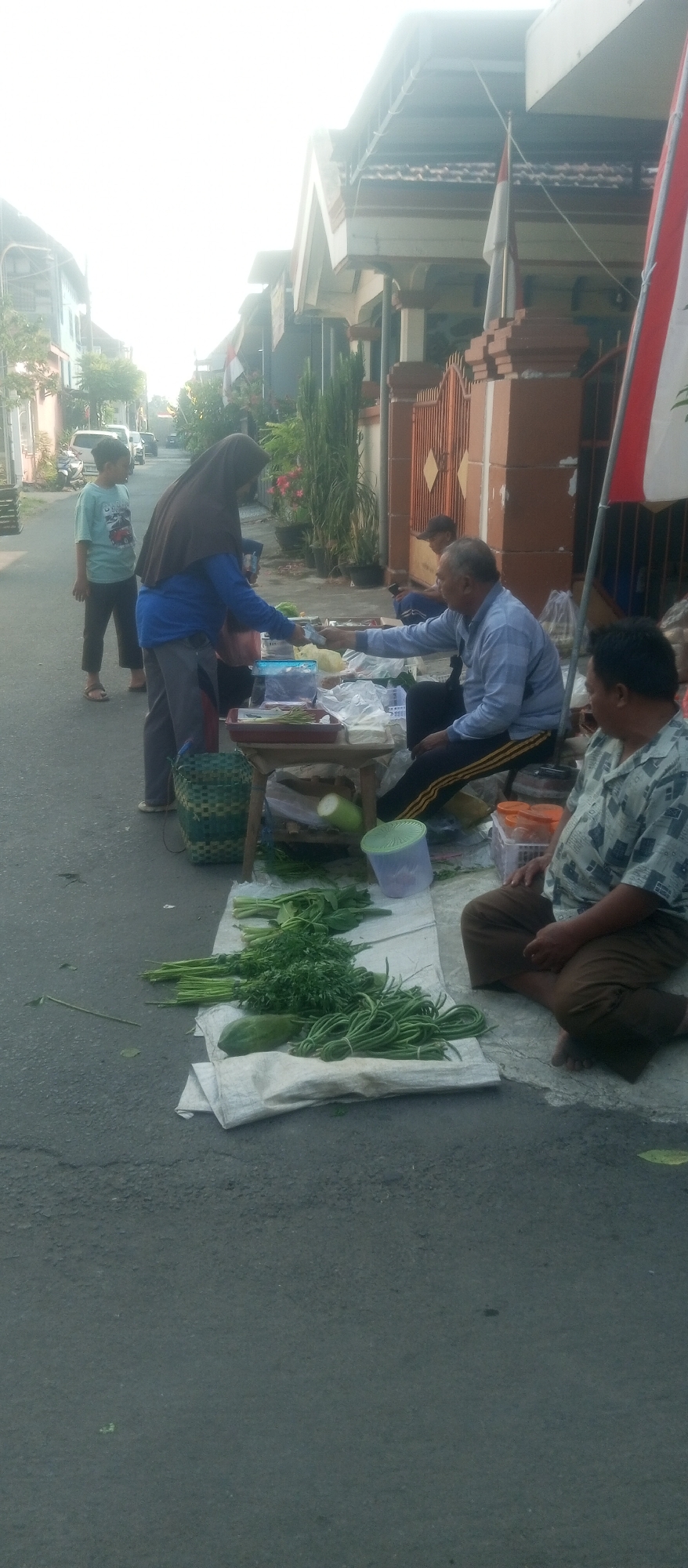 In an outdoor scene in the city of Kediri, Indonesia, likely a street market, a vendor in a blue shirt and headscarf is interacting with a customer. The vendor is standing and handing over some items, possibly food, to a man who is seated. To the left, a young boy in a light blue t-shirt and dark pants stands on the side of the road. In the foreground, a variety of fresh vegetables, including long beans, leafy greens, and a papaya, are displayed on a white tarp spread on the asphalt. Behind the seated customer, another man is sitting and observing. A small wooden table with various items, including plastic containers, is also present. The background shows a residential street with houses and vegetation, and a national flag is visible on a pole. The overall lighting suggests it is daytime, possibly morning or late afternoon.