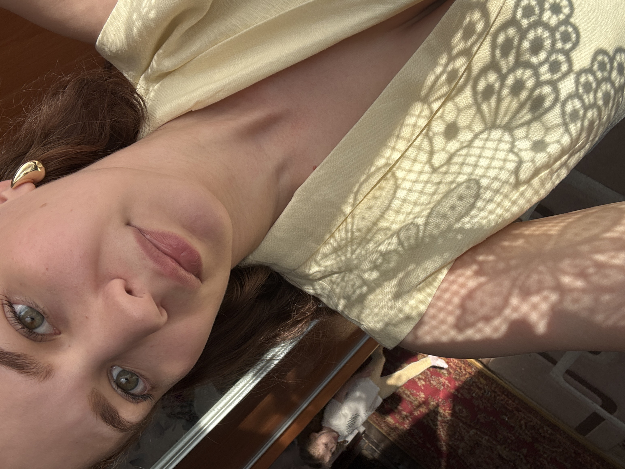In a close-up selfie, a woman with light brown hair and green eyes is positioned in the foreground, looking directly at the camera. She is wearing a light yellow top and a distinctive gold, horn-shaped earring on her right ear. The sunlight casts a decorative shadow pattern across her clothing. In the background, visible through a doorway or window, a young girl wearing a white t-shirt and yellow leggings is walking on a red patterned rug. The environment suggests an indoor setting with natural light entering from the right. The location is identified as Nizhnyaya Gar', Russia.