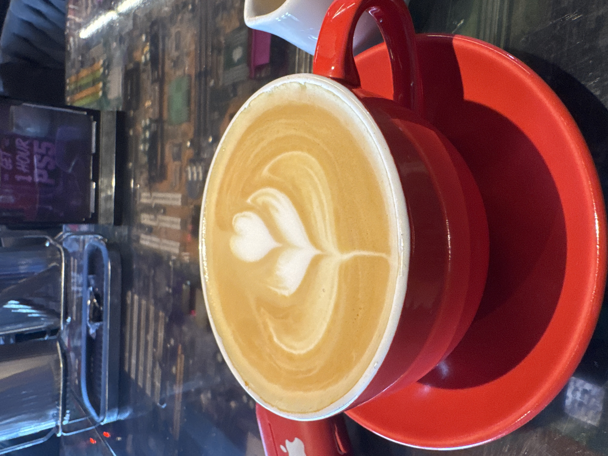 A red coffee cup and saucer are filled with a latte topped with latte art resembling a heart and a swirl. The cup and saucer are a vibrant red, and the latte is a creamy brown with a white foam art. In the background, there is a sign that reads "GET 1 HOUR PS5" and other blurry objects that appear to be in a cafe or gaming establishment. The table surface is reflective and appears to be black with some subtle patterns. To the left of the cup, a red object that may be an Apple product is visible. To the right, a small white creamer is also present. The overall scene is a close-up shot focusing on the coffee and its artistic presentation. The location of Kuta Selatan, Indonesia, is not discernible from the image itself.