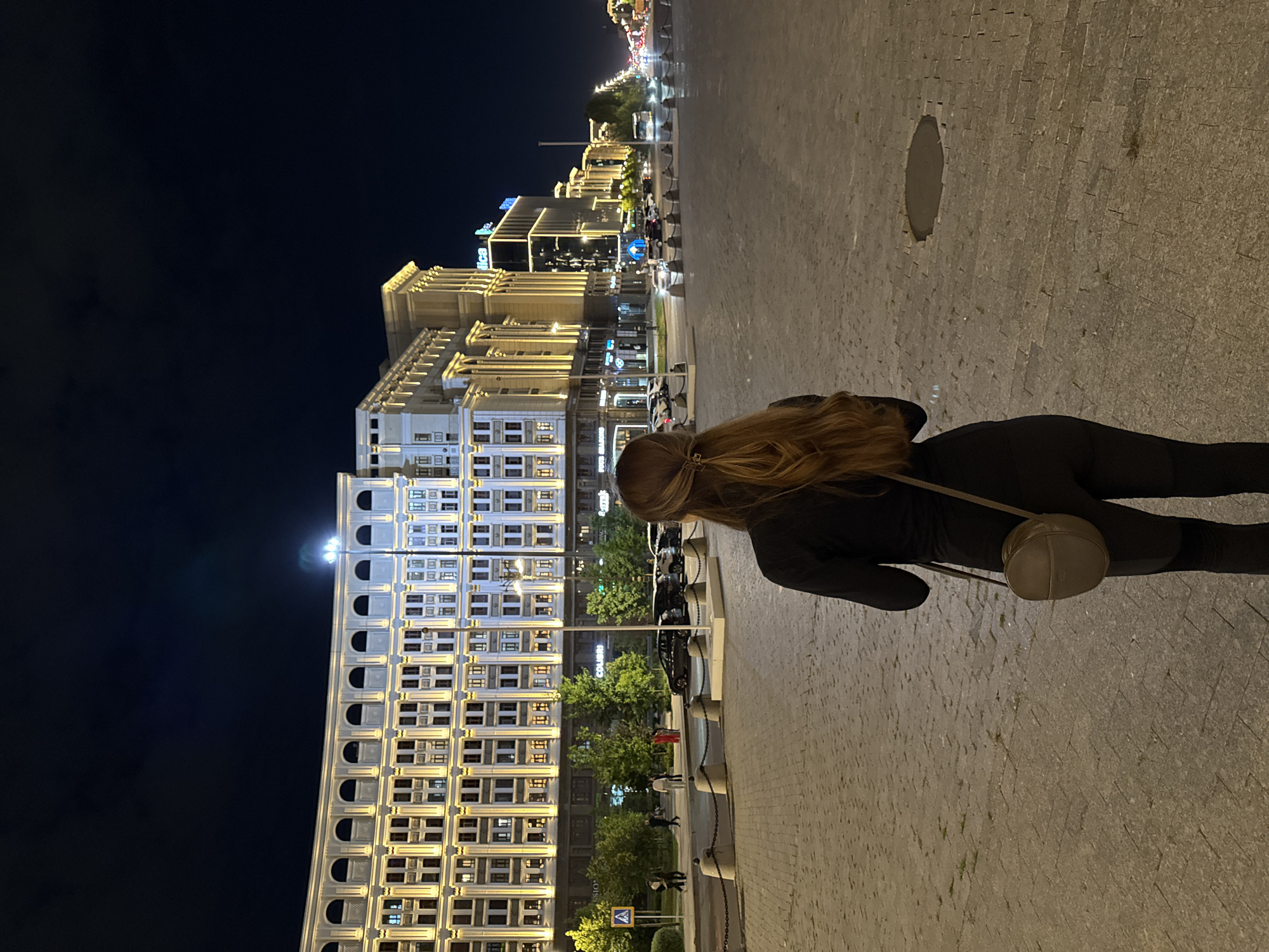 A woman with long, light brown hair tied back stands in a large, open plaza paved with cobblestones at night. She is wearing a dark, form-fitting jumpsuit and a small, round, light brown crossbody bag. Her back is to the camera, and she is looking towards a grand, ornate building illuminated with warm yellow lights. The building has multiple floors with arched windows and decorative architectural details. Trees line the street in front of the building, and cars are parked and driving in the background. Beyond the initial building, other structures with illuminated windows and signage stretch along a wide street, suggesting a city environment. The sky is dark, indicating nighttime. The overall scene depicts an urban setting at night, with the woman as the primary subject in the foreground, observing the illuminated cityscape.