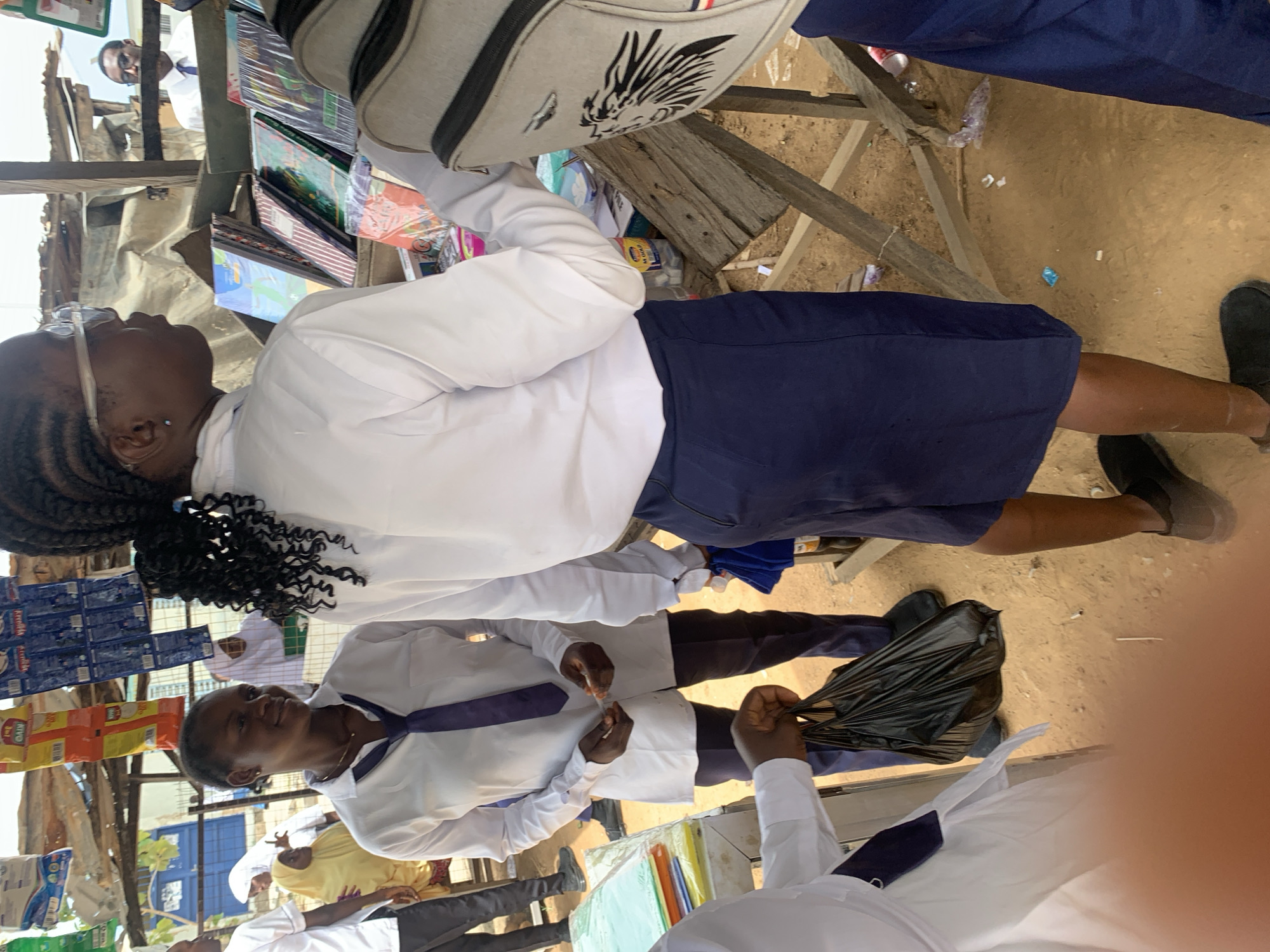 In Keffi, Nigeria, the image shows a group of people, most likely students in school uniforms, near a roadside stand. The students appear to be making purchases, possibly of school supplies. The stand has various items on display, including what appears to be food items, notebooks, and other goods. The ground is dirt, and there are other people in the background. The weather appears to be clear and bright, possibly during daylight hours.