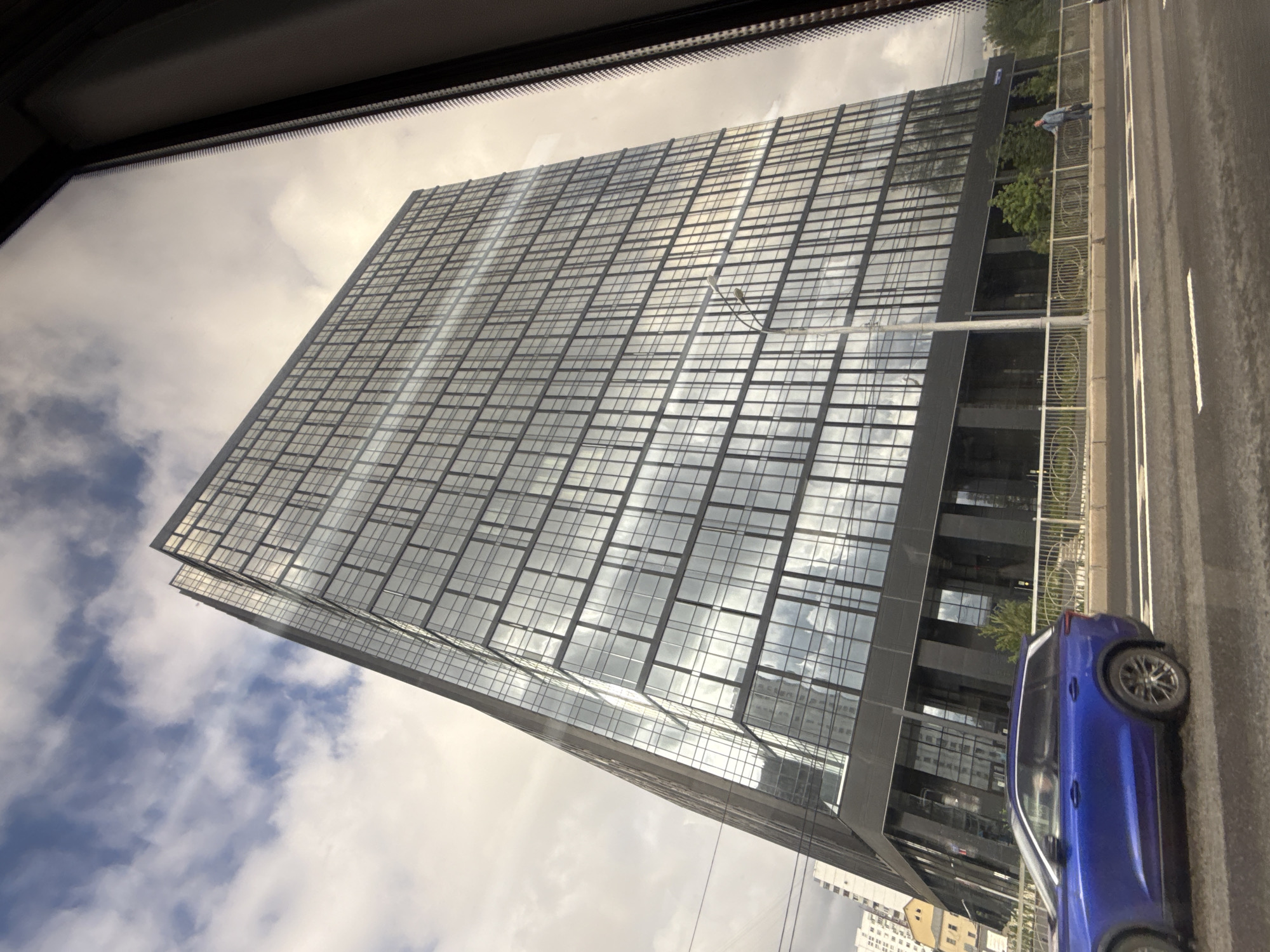 Here's a detailed description of the image:
The image shows a modern, high-rise building with a predominantly glass facade, reflecting the sky and clouds. The architecture features a grid-like pattern of windows and a sleek, minimalist design. A blue car is parked on the right side of the frame, next to a road. Beside the road is a pedestrian area with a railing. A person is visible standing along the railing. The photo is taken from inside a vehicle, with the car's interior visible in the top left corner of the frame. The angle suggests the building is very tall, as it dominates the view upwards. Based on the architecture and available context clues, the image may be from Moscow, Russia.
