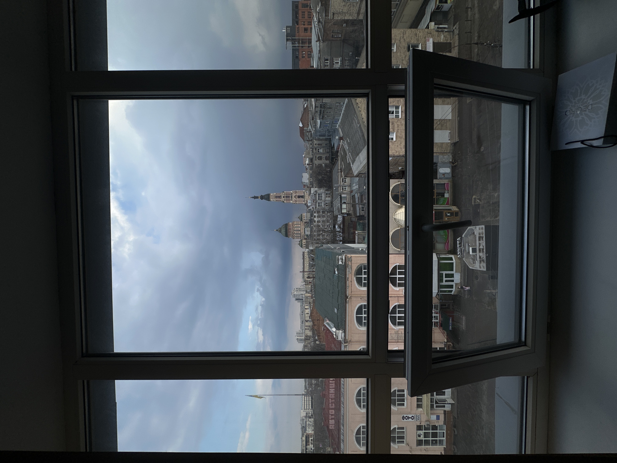 The image shows a view from a window of a building in Kharkiv, Ukraine. An open window pane is on the right side of the frame. Outside the window is a cityscape. The Dormition Cathedral and its bell tower are visible in the distance. Other buildings, including some with arched windows, are also visible. The sky is overcast with dark clouds. A Ukrainian flag on a tall pole is visible. A shopping bag rests on the windowsill to the right.