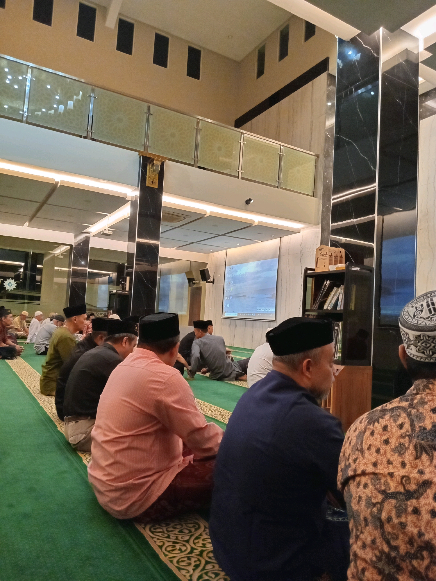 In Randuagung, Indonesia, a group of men are seated on a green carpet in what appears to be a mosque. They are facing a large screen displaying an image of a sky with clouds, possibly a presentation or lecture. The men are dressed in traditional Islamic attire, mostly wearing peci (caps) and sarongs or trousers. Some are looking at the screen, while others are engaged in conversation or prayer. The interior of the mosque features modern architecture with marble columns and glass railings on an upper level. A bookshelf containing books and other items is visible next to one of the columns. The lighting is soft and ambient, creating a peaceful atmosphere. The overall scene suggests a religious gathering or educational event.