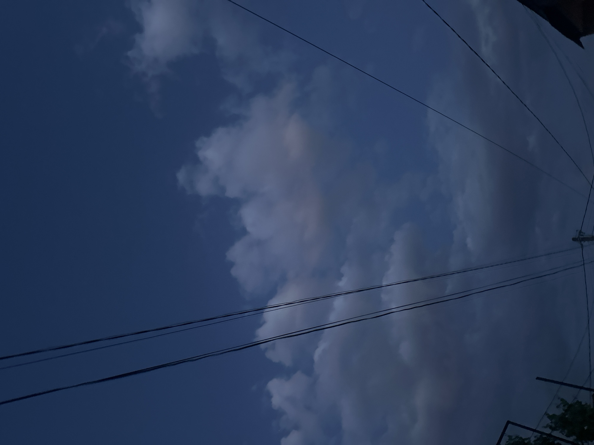 The image shows a dark blue sky partially covered by clouds. Several power lines run diagonally across the frame. A portion of a building is visible in the lower right corner, along with some greenery. The location is likely a residential area in Yerevan, Armenia, although the specific neighborhood cannot be determined from the image alone. There are no people visible.