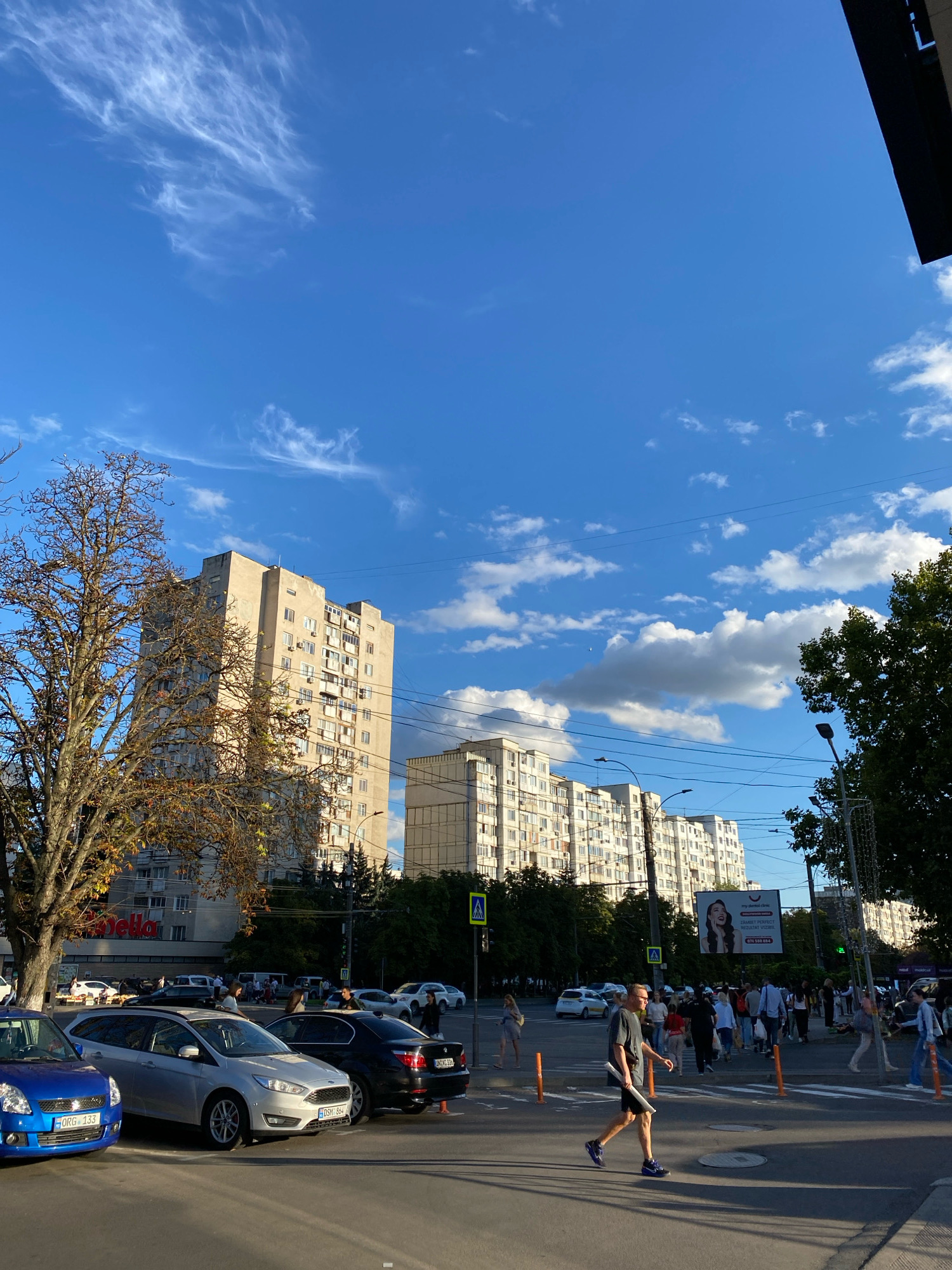 The image depicts a street scene in Chișinău, Moldova, on a sunny day with scattered clouds. A tree with autumn foliage stands prominently in the foreground on the left, casting a shadow. Behind the tree, several multi-story apartment buildings are visible, characteristic of Soviet-era architecture. The street is lined with parked cars and a pedestrian crossing is active, with several people walking. A man in a gray t-shirt and black shorts is crossing the street, carrying a white tube. In the background, there is a billboard advertising dental services, and more apartment buildings stretch into the distance. Power lines are visible overhead, strung between utility poles. The overall impression is a typical urban environment in Eastern Europe during the daytime.