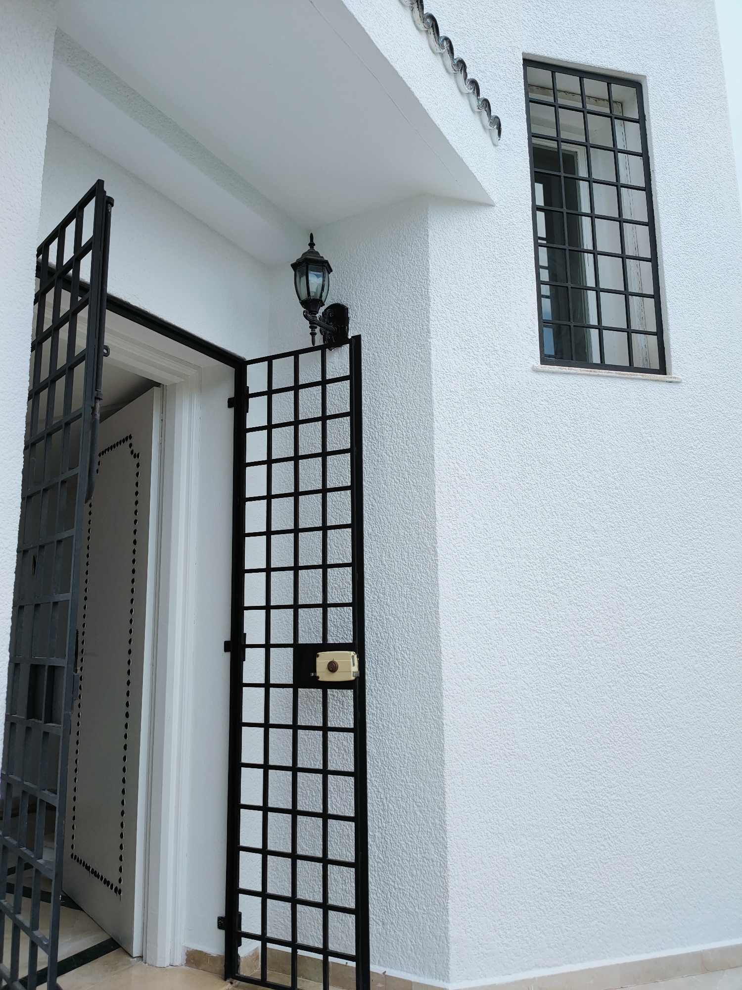 The image shows an exterior view of a white, textured building. A black metal gate with a grid pattern is open, revealing a white door with decorative perforations. A black wall lamp is mounted on the wall above the gate. To the right of the gate, a tall, narrow window with a black metal grid is visible. The scene appears to be in the city of Mrezga, Tunisia, suggested by the architectural style and textured stucco walls common in the region.