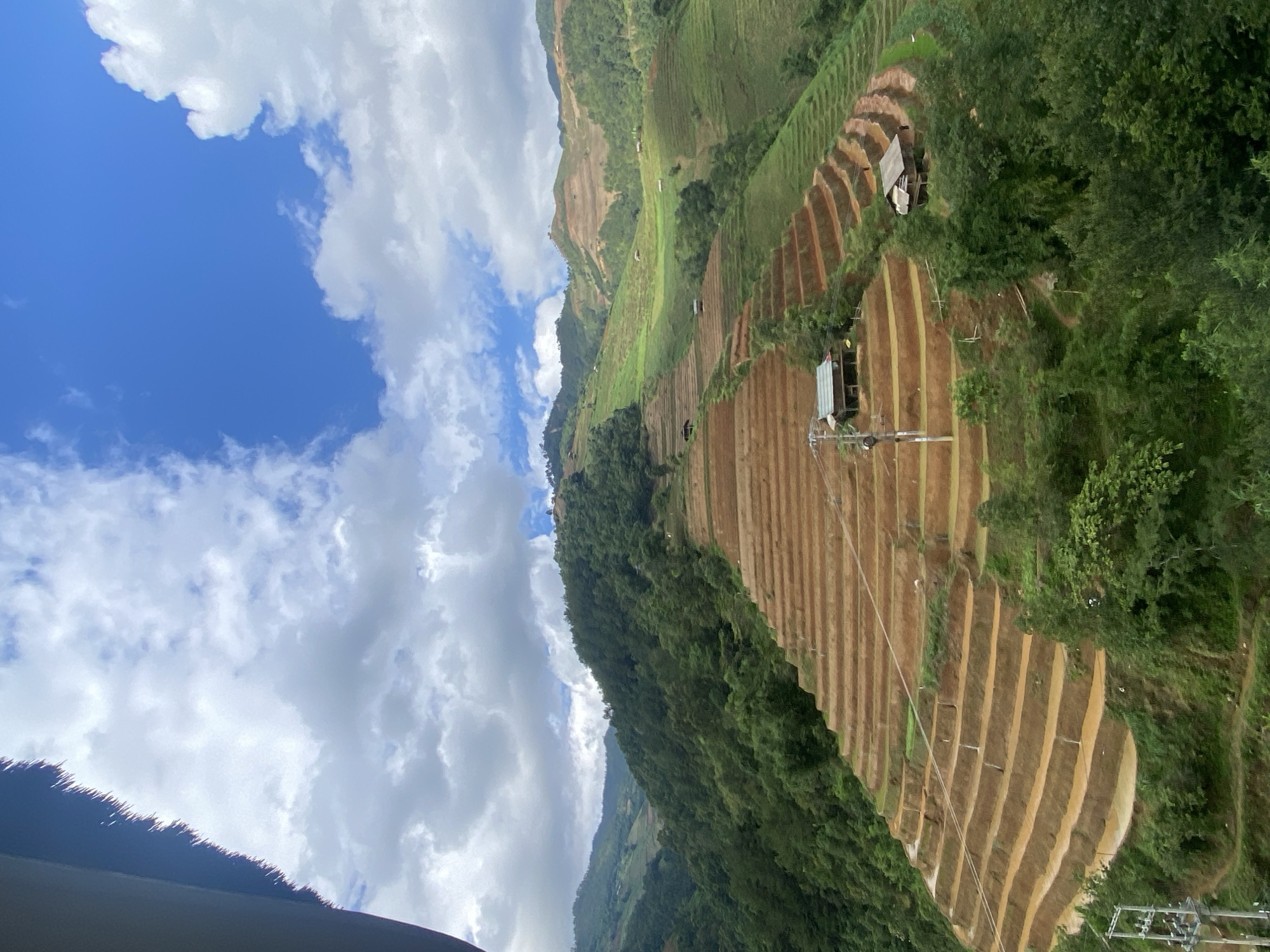 This forensic description details an expansive mountainous landscape, identified as Mù Cang Chải, Vietnam, characterized by extensive agricultural terracing under a partially cloudy sky.
The upper portion of the image displays a sky with clear blue patches, predominantly in the upper right quadrant, contrasting with large, white and grey cumulus clouds that occupy the remaining sky. A dark, out-of-focus element, resembling the underside of a thatched roof or eave, partially intrudes from the top-left corner.
The dominant visual elements are the numerous rice terraces meticulously carved into the steep slopes of multiple mountains. These terraces exhibit varying states: a significant portion, particularly in the mid-left and foreground, shows reddish-brown earth, indicative of recently tilled or fallow land. In contrast, sections on the right side and in the upper mid-ground are covered in vibrant green vegetation, consistent with active rice cultivation. The terraces follow the contours of the hillsides, forming intricate, curvilinear patterns.
Beyond the cultivated areas, the mountains are densely forested with various shades of green trees and undergrowth, receding into the distance where light and shadow differentiate their forms.
Several man-made structures are integrated into the landscape. Two prominent, small, rustic huts with dark roofs and light-colored walls are situated on different terraced levels: one in the mid-left on a lower terrace, and another further to the right and slightly lower. Additional, smaller, less distinct huts are visible scattered across the distant green terraces. In the lower-left foreground, a utility pole with multiple crossarms and associated power lines is visible, indicating localized infrastructure.
The scene is uniformly illuminated by natural daylight, casting subtle shadows that define the terrain. No human or animal figures are present within the frame.