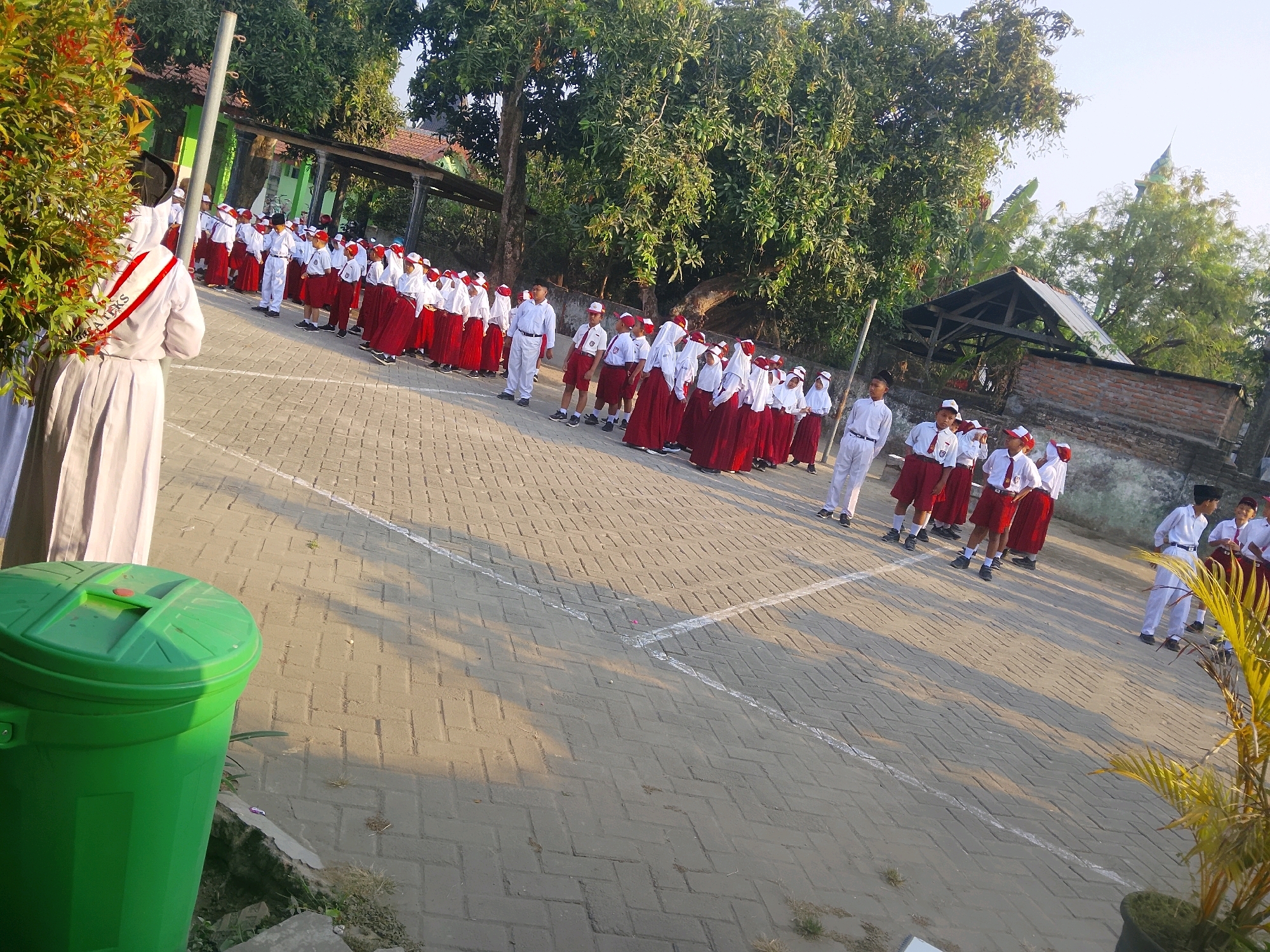In the city of Kepuh, Indonesia, an outdoor scene unfolds with a group of students, appearing to be in uniform, standing in rows on a paved area. The students are dressed in white shirts and red skirts or pants, with some wearing white head coverings. In the foreground, a large green trash can is visible. A woman, possibly a teacher or supervisor, stands to the left, facing away from the camera. She wears a white garment with a red and white sash. The background features trees and a building with a tiled roof. The lighting suggests it is either early morning or late afternoon, with shadows cast on the paved ground.