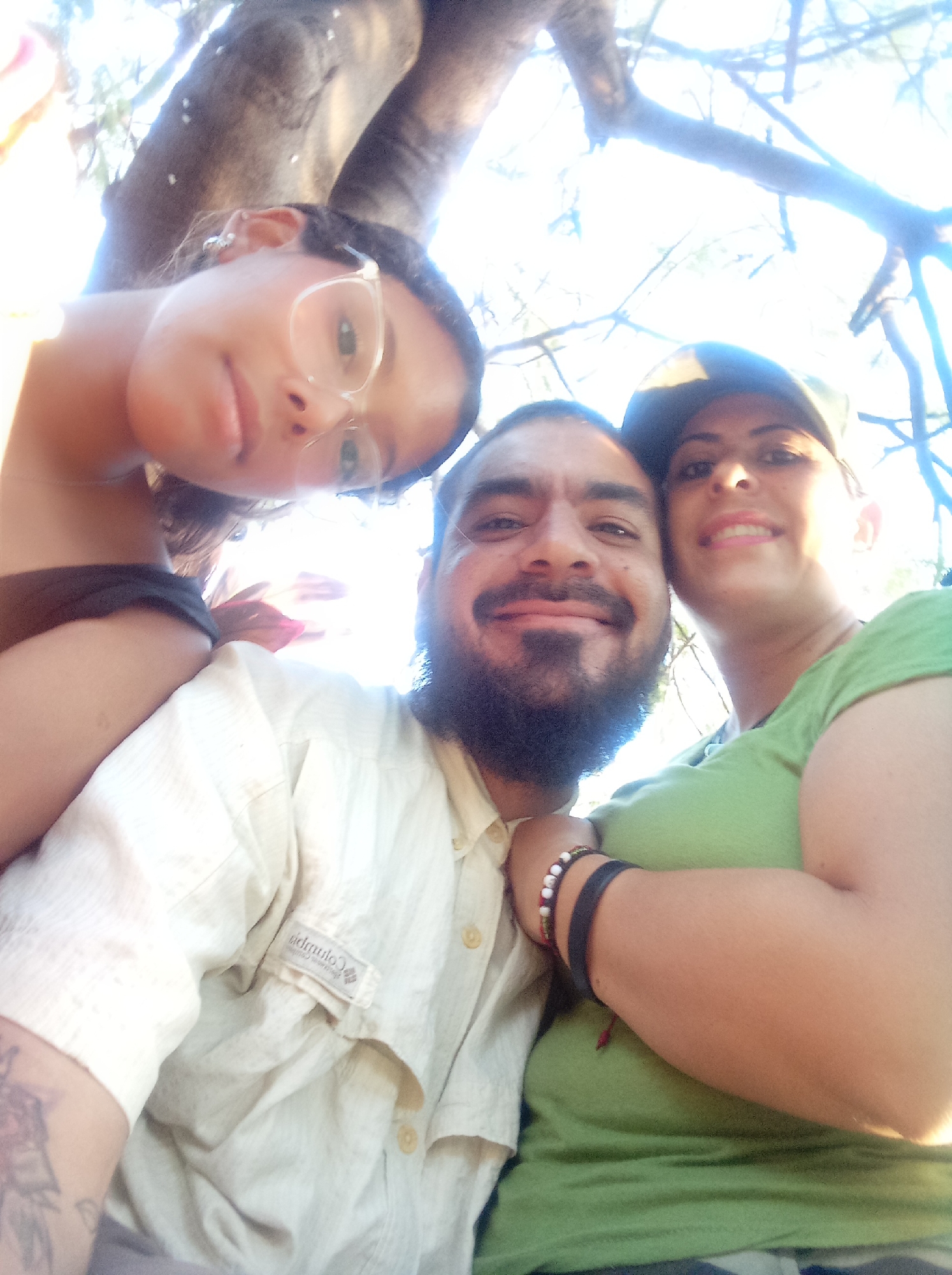 This is a brightly lit, low-angle selfie of three people smiling happily outdoors in Guarenas, Venezuela. The strong, direct sunlight and lens flare suggest it is daytime, likely a sunny morning or afternoon.
On the left, a young woman with dark hair and clear-rimmed glasses looks gently at the camera. She wears a dark top and appears to be leaning over the central figure. In the middle, a man with a dark beard and mustache beams at the camera. He is wearing a light-colored, short-sleeved button-up shirt, which clearly displays a "Columbia" brand tag on the chest pocket. His right wrist is adorned with a black band and a multicolored beaded bracelet. To the right, a woman with dark hair and a cap smiles warmly. She is wearing a light green t-shirt, and her arm is around the man in the center.
The background features blurred tree branches and bright sky, indicating an outdoor setting. All three individuals appear happy and relaxed, enjoying their time together.