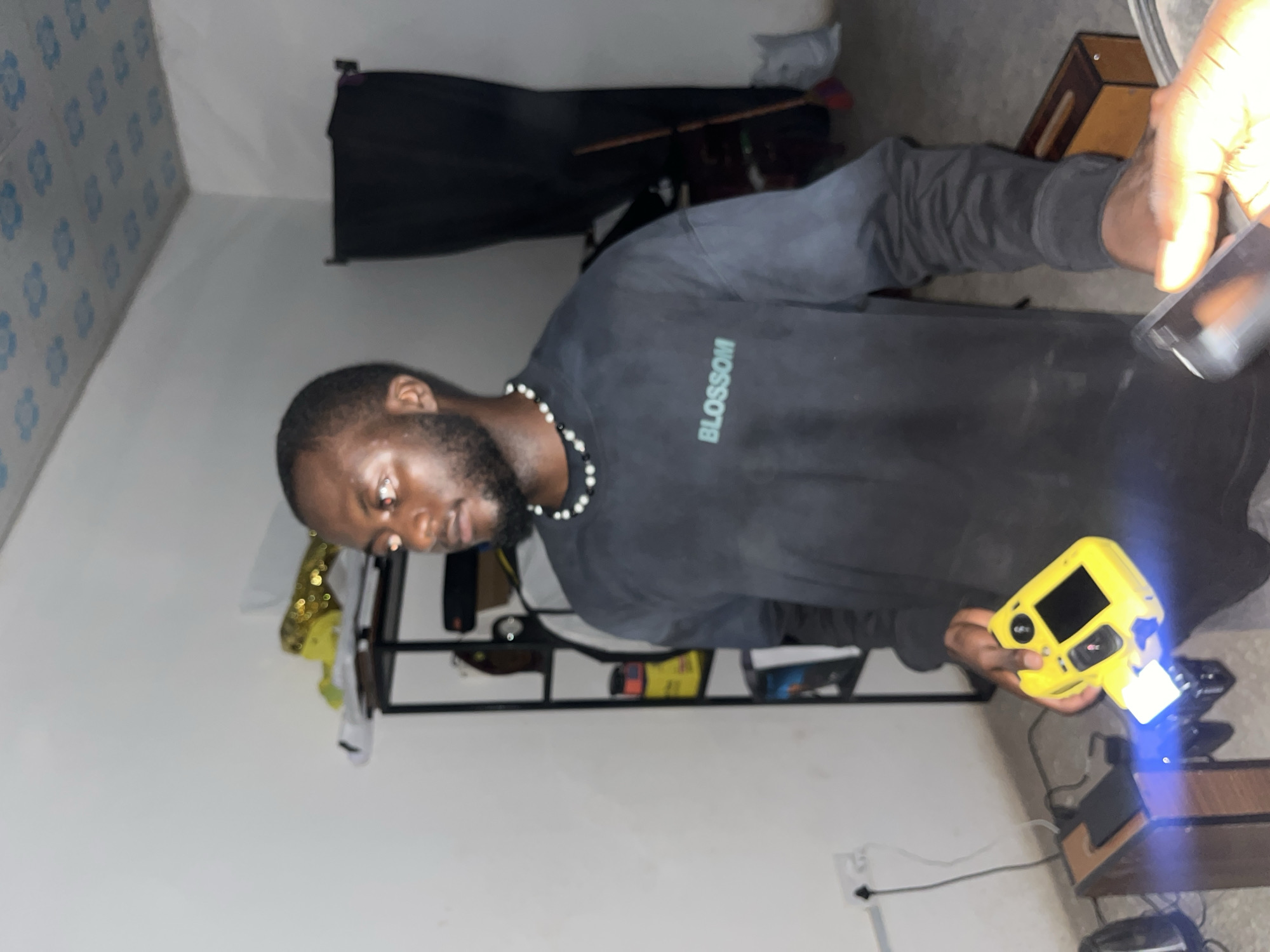 In a dimly lit room, likely in Gura Top, Nigeria, a young Black man with a beard stands facing right, holding a yellow electronic device with a bright blue light emanating from it. He wears a black long-sleeved shirt with the word "BLOSSOM" printed in teal on the chest and a beaded necklace. To his left, a black shelf unit is partially visible, containing various items including yellow and black containers. Behind him, a large black fabric is draped, possibly a backdrop or curtain, and a broom leans against it. The room has white walls and a patterned ceiling with blue floral designs. On the floor to the man's left, a dark brown wooden object with electronics is present. His right hand is near a black rectangular object, possibly a phone, which is out of focus.