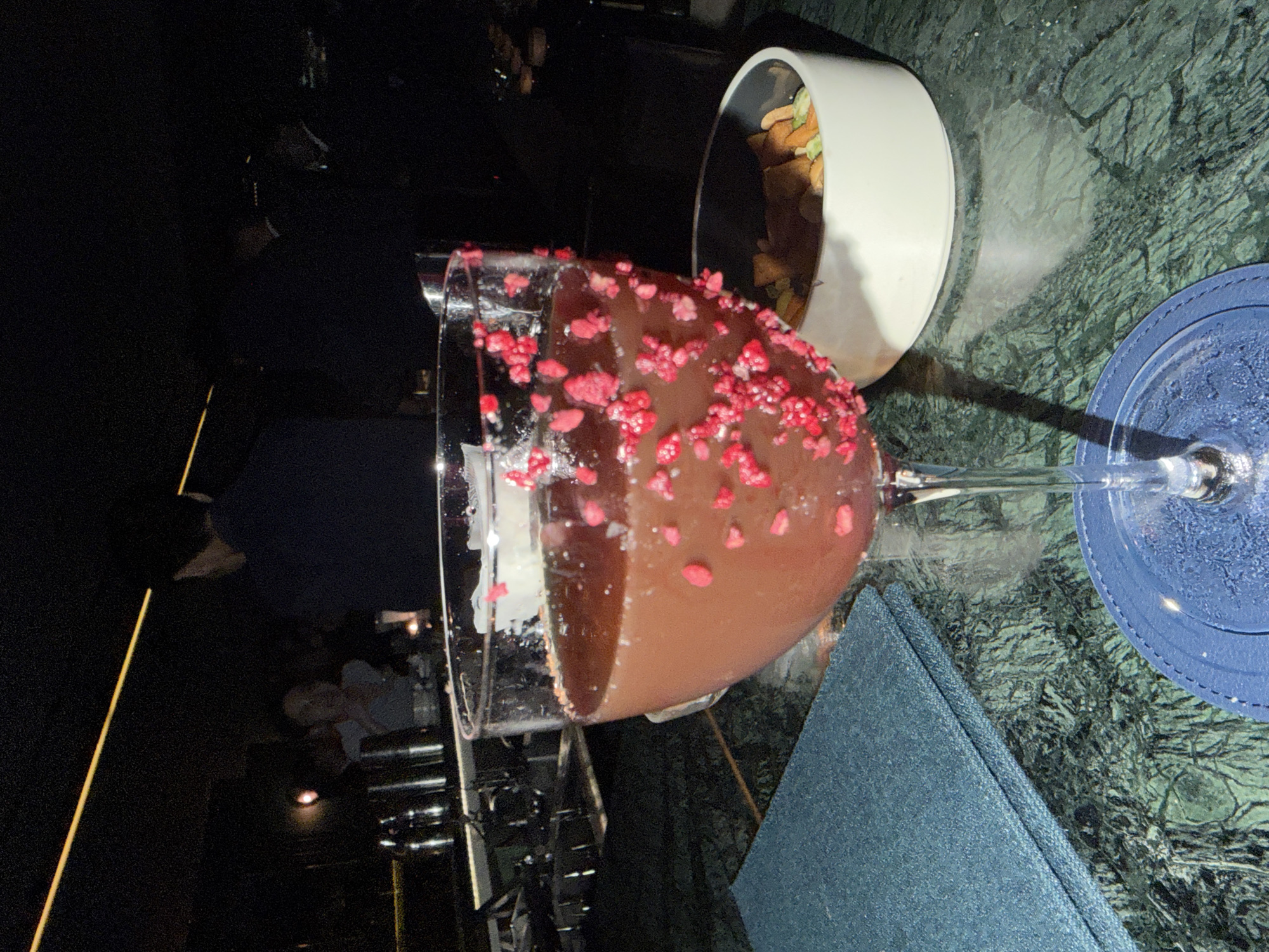The image shows a cocktail in a large coupe glass, rimmed with small red berries, sitting on a dark green marble table with a blue napkin and matching coaster. Beside the glass is a white bowl containing snacks. In the background, people are seated at a dimly lit bar or restaurant. The image was taken in Bangkok, Thailand.
