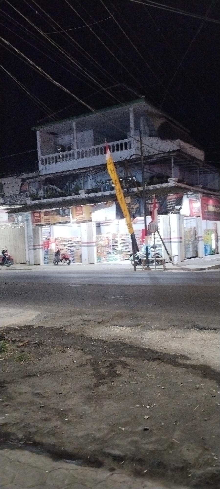 This is a nighttime photograph of a two-story building with a covered walkway in front, possibly a storefront or shop, on a street in Jombang, Indonesia. The building has a white balustrade on the second-floor balcony. A tall, yellow banner with Indonesian text and two small Indonesian flags are visible attached to a pole in front of the building. Several motorcycles are parked on the street or being ridden. The street surface appears to be asphalt and is somewhat uneven with some patches of darker material. Overhead, numerous wires are strung across the scene. The lighting suggests artificial illumination from the building's interior and possibly streetlights.